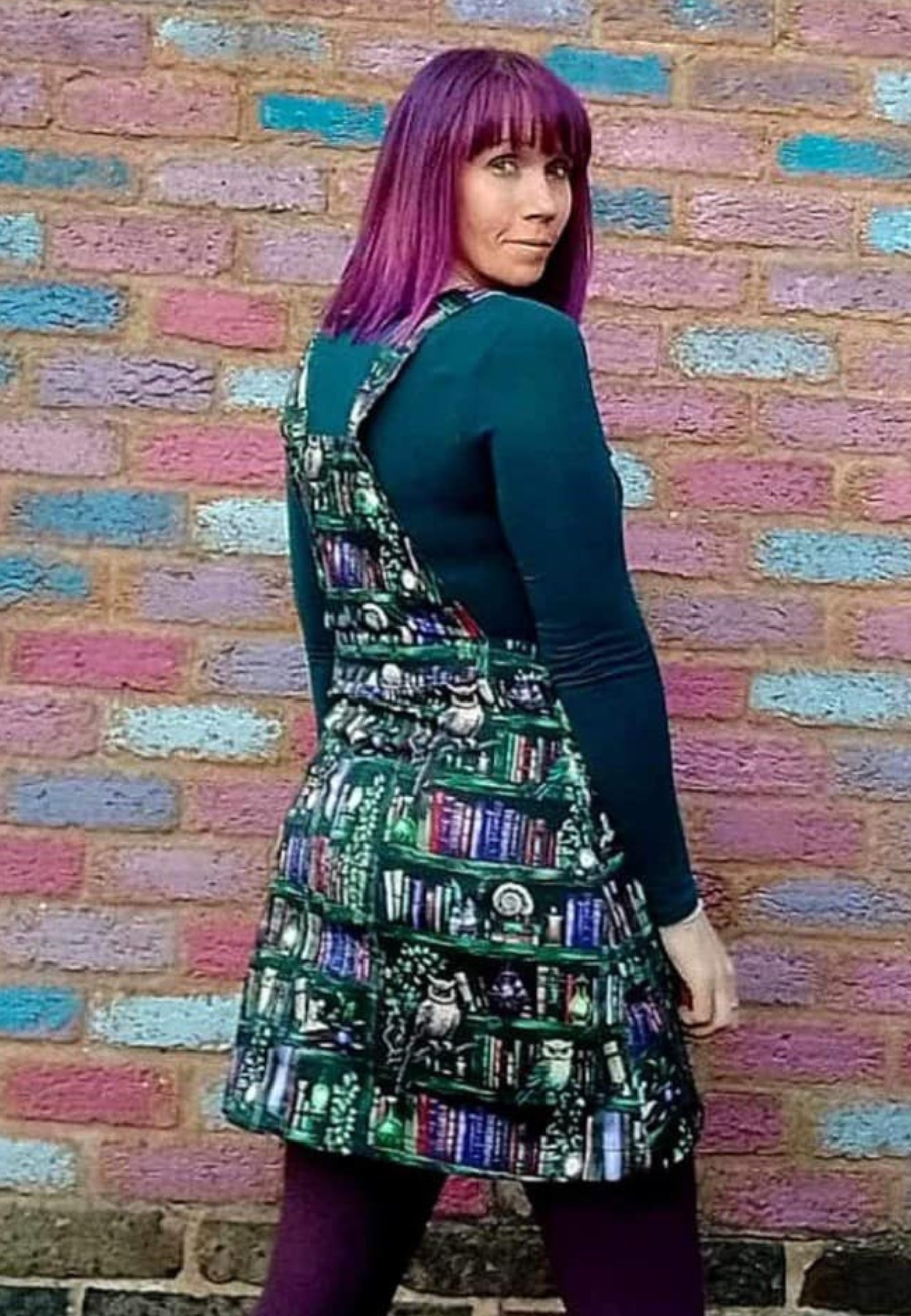A woman wearing a green pinafore dress with a unique bookcase and owls print, standing in front of a colorful brick wall. Back view of pinafore. 