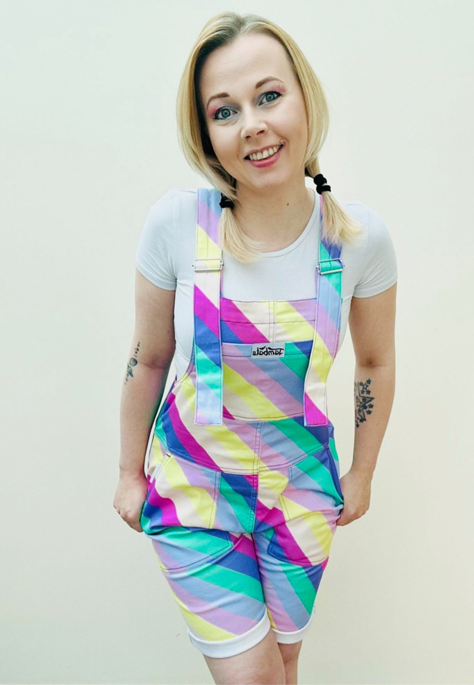 A person wearing bright multicolored striped shorts dungarees with a chest pocket, standing against a plain background. Bright pastel striped dungaree shorts. Retro style. 