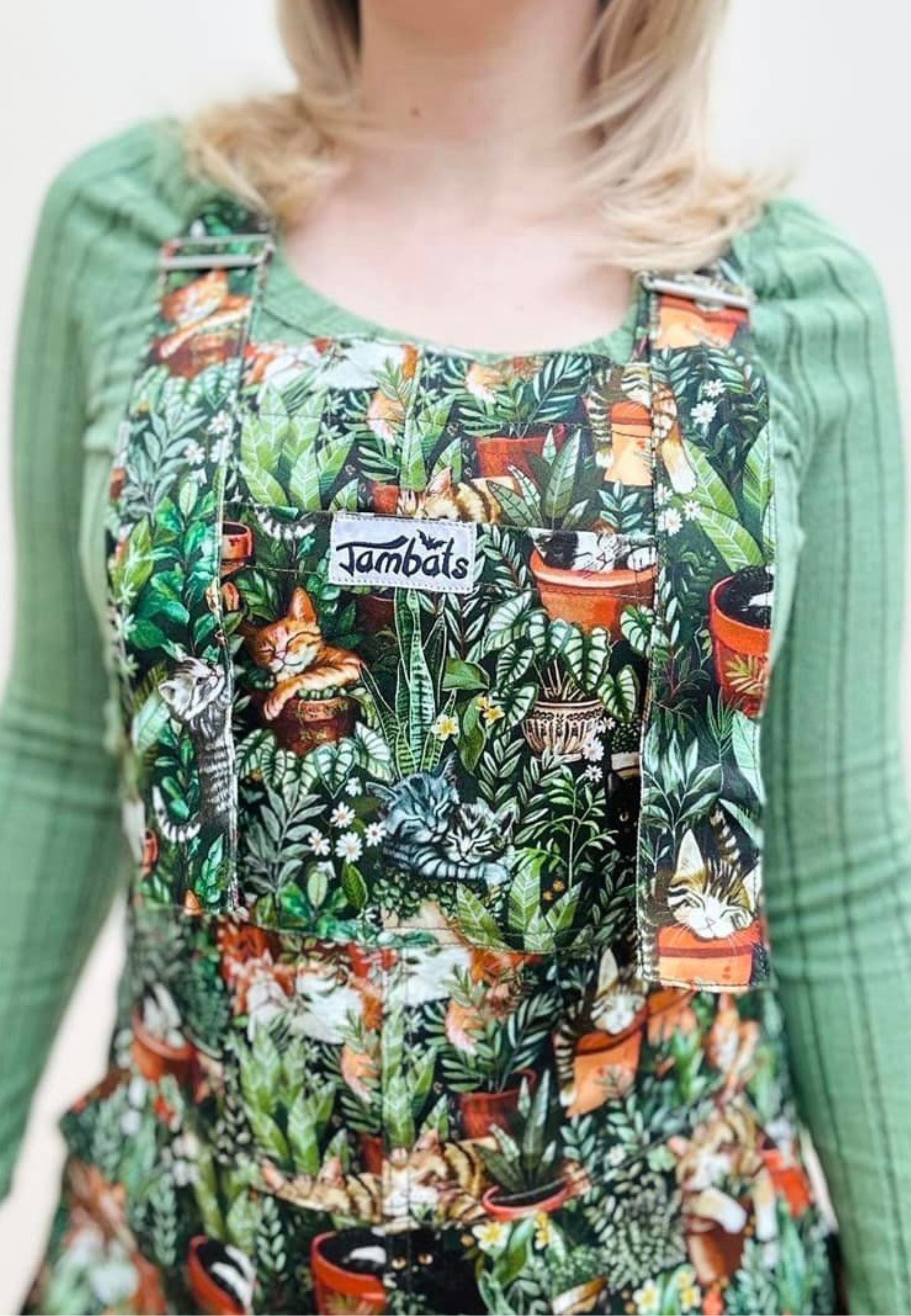 A woman wearing green Cat Nap dungarees with a pattern of various sleeping cats among houseplants. The dungarees have chest, hip, and bum pockets and feature a slider buckle fastener.
