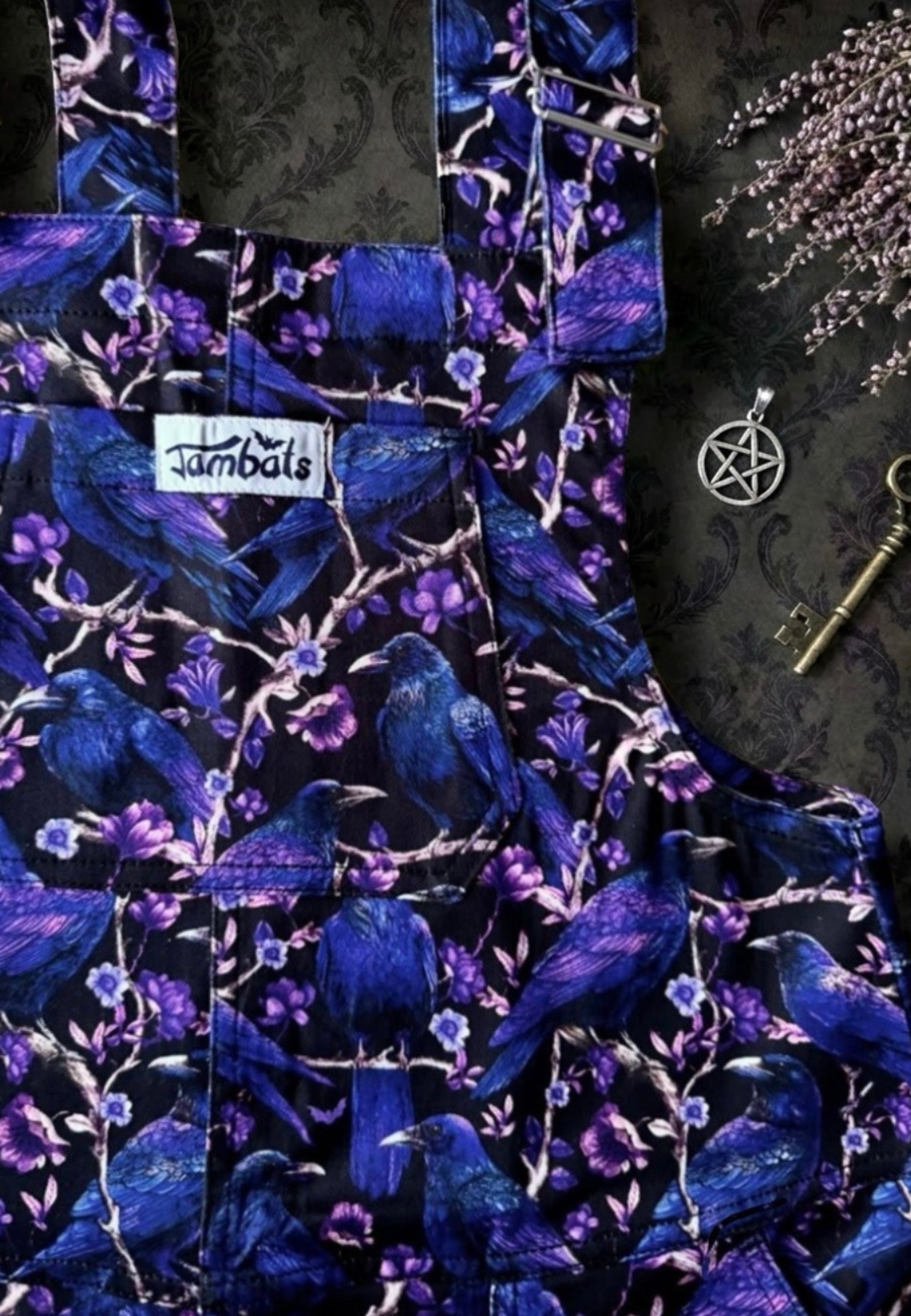 Flat lay of beautiful corvid print dungarees. Print is of deep purple and blue crows and ravens perched on blooming branches on a black background. Jambats dungarees