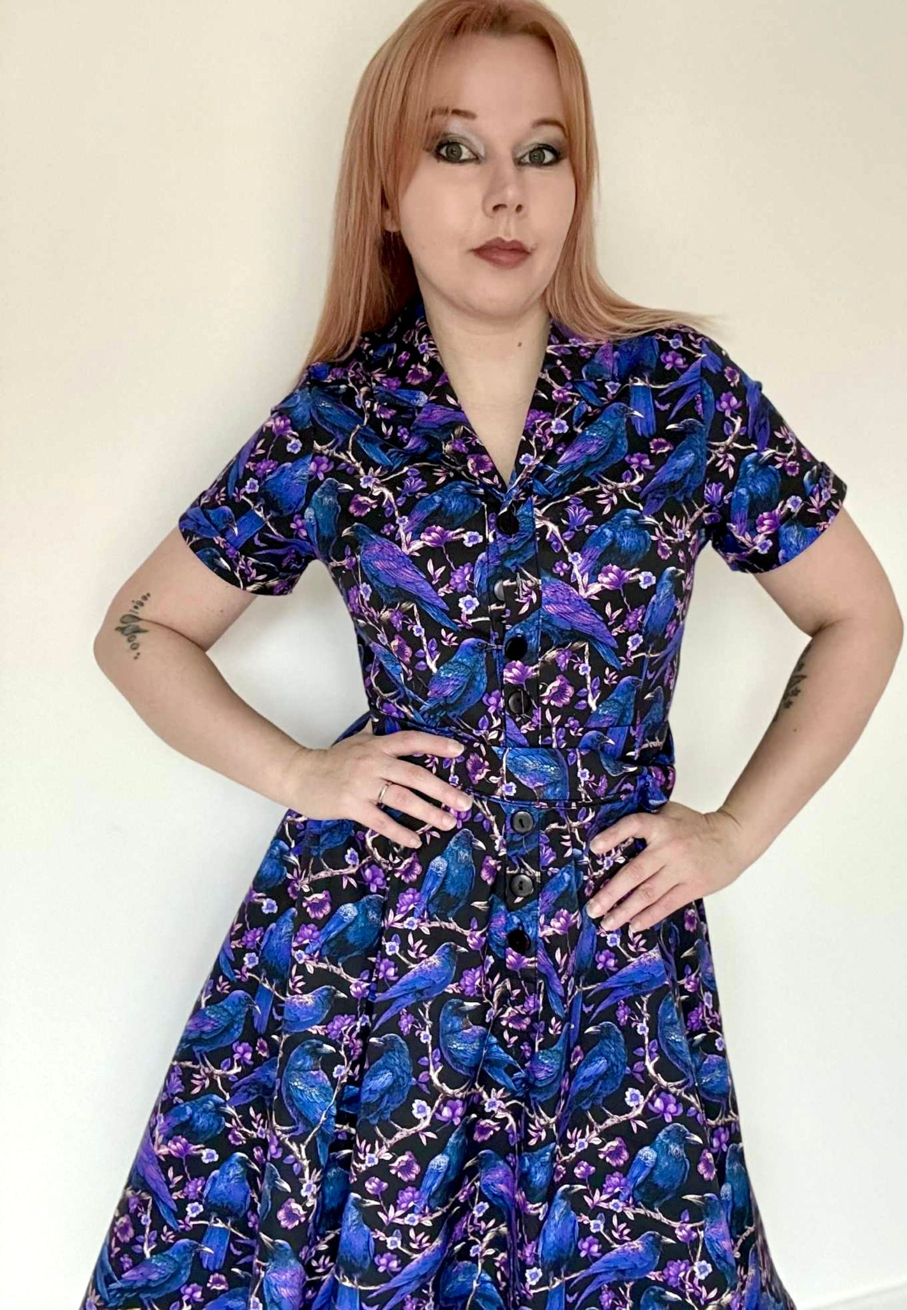 Woman wearing a beautiful Corvid print dress. Dress is black with blue and purple crows and raven perched amongst branches. Dress is a retro shirt style, full skirt and button up blouse. Soft goth style. 