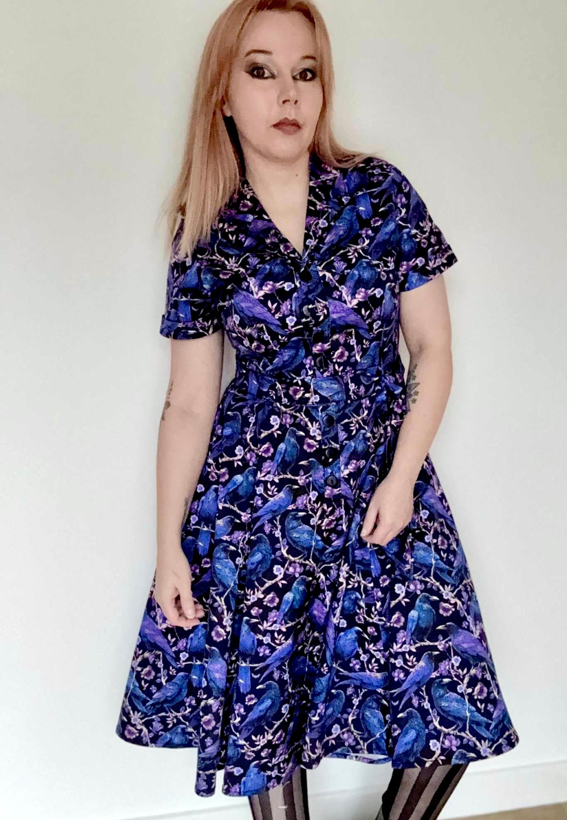 Woman wearing a beautiful Corvid print dress. Dress is black with blue and purple crows and raven perched amongst branches. Dress is a retro shirt style, full skirt and button up blouse. Soft goth style