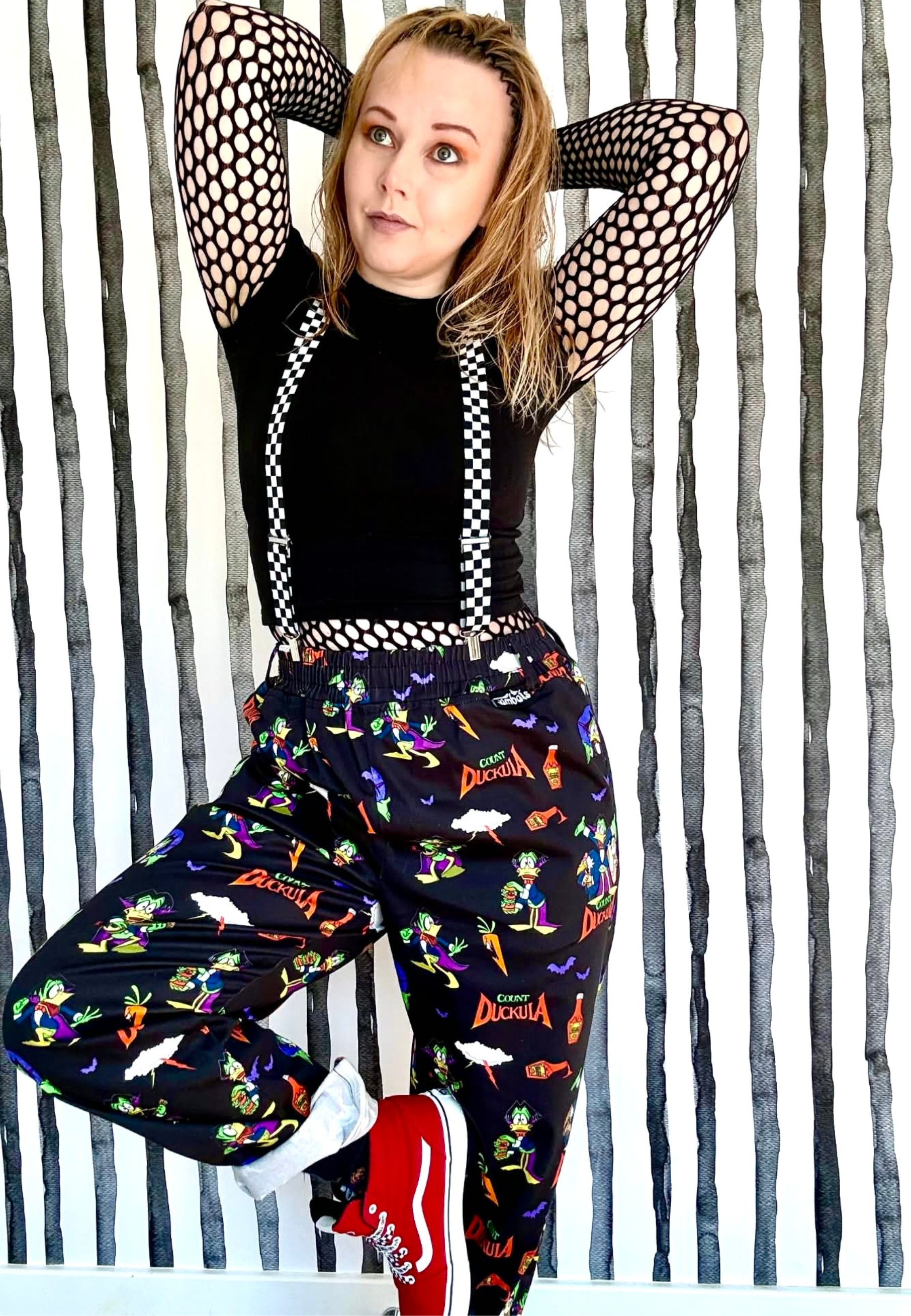 Person wearing a black top with checkered straps and Count Duckula trousers against a striped wallpaper background