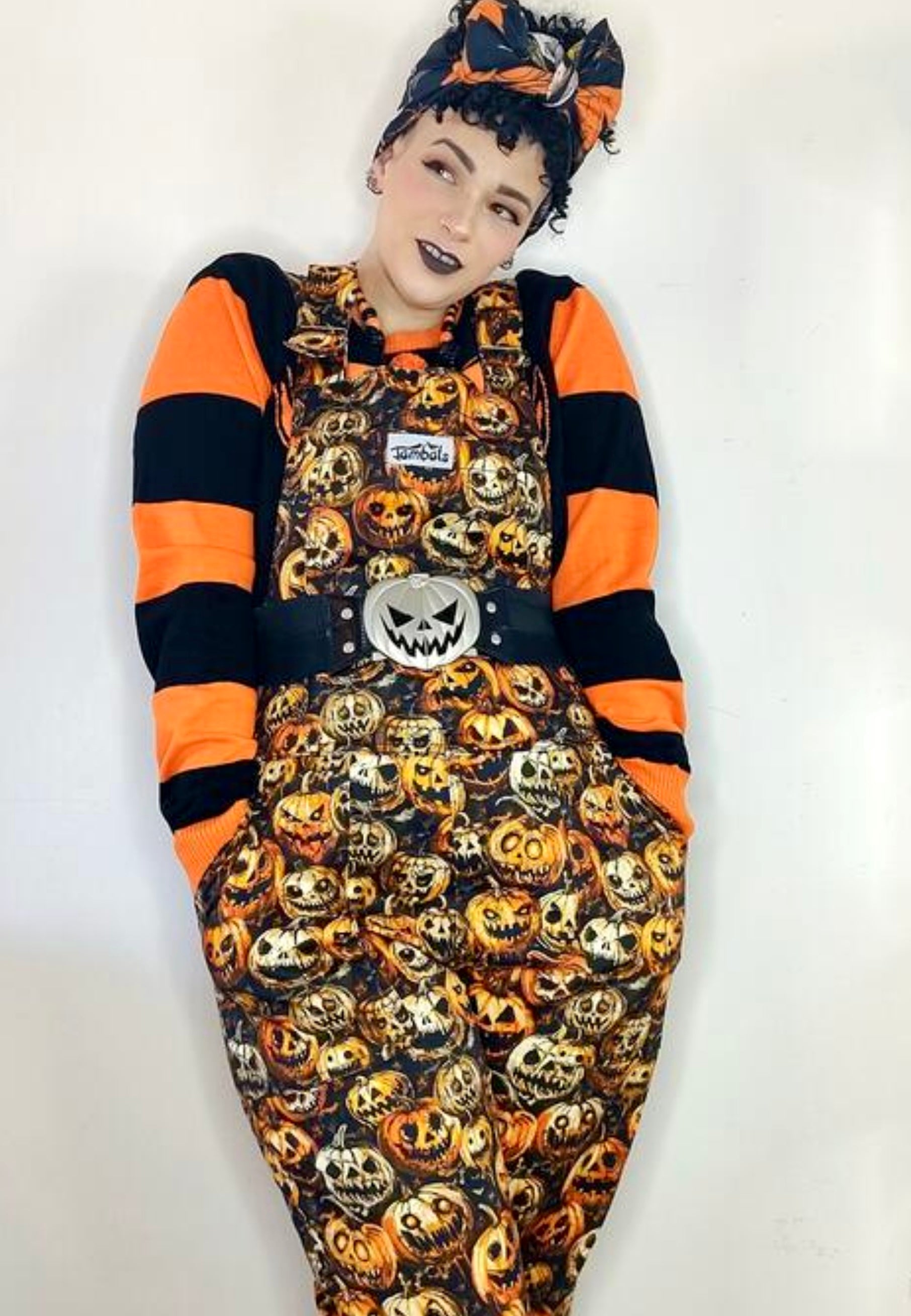 Model wearing Halloween themed dungarees overalls with illustrated creepy pumpkins print. Model is also wearing orange and black striped jumper and hair tie. Quriky retro spooky Hallween style