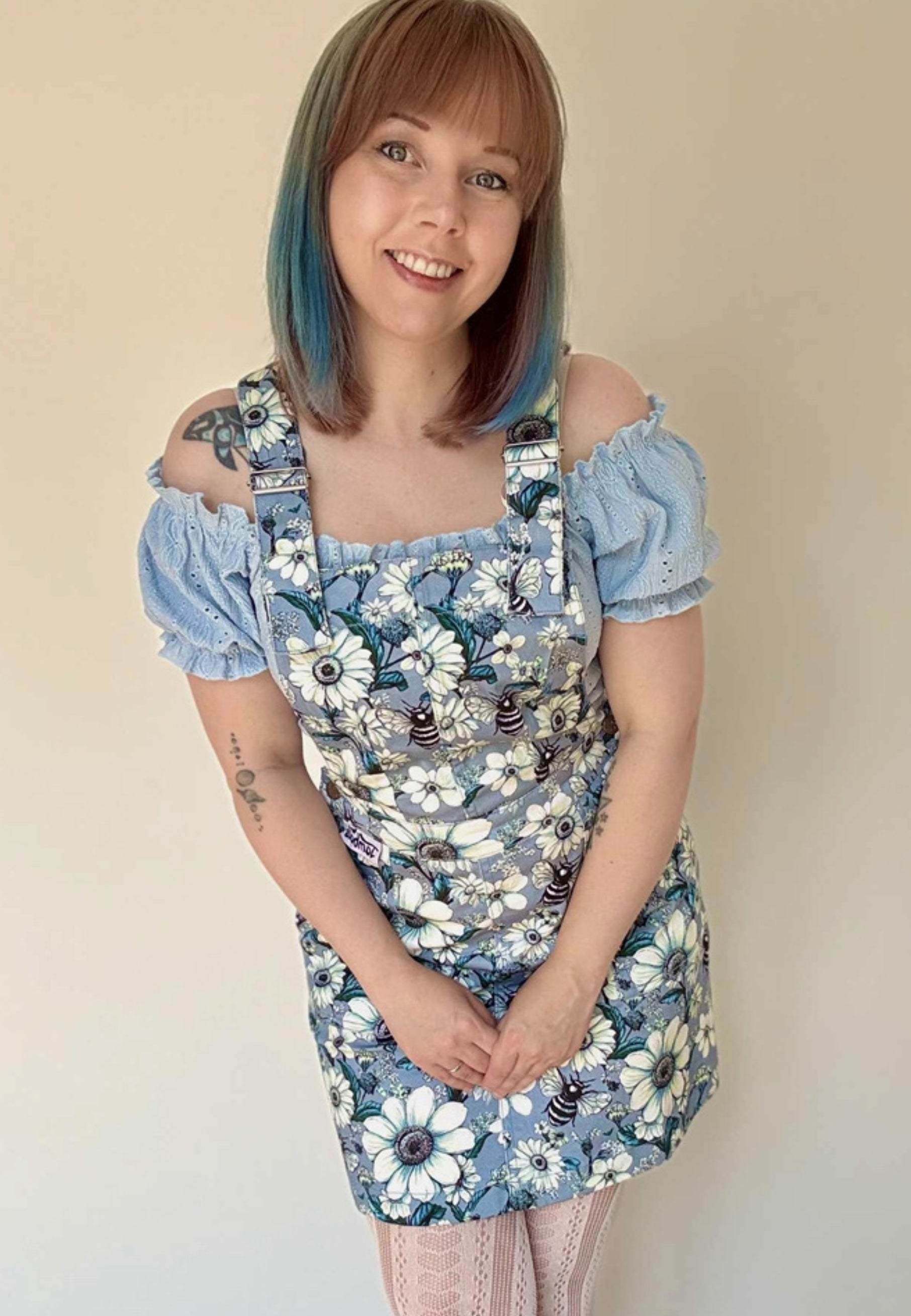 Woman wearing a floral pinafore dress with a print of daisies and bees,over a puff sleeve shoulder top against a beige background. Cottagecore style