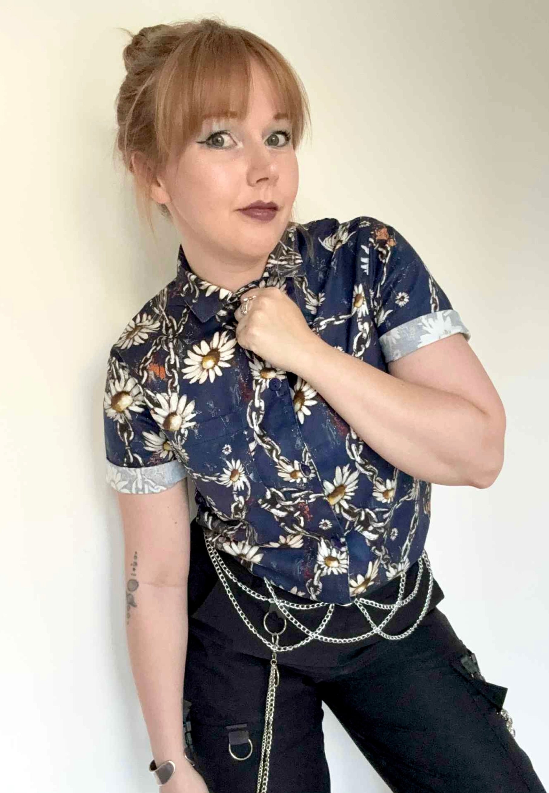 Woman wearing a deep blue shirt  with chains and daisies and dark pants against a plain background. Grungey floral style print. 