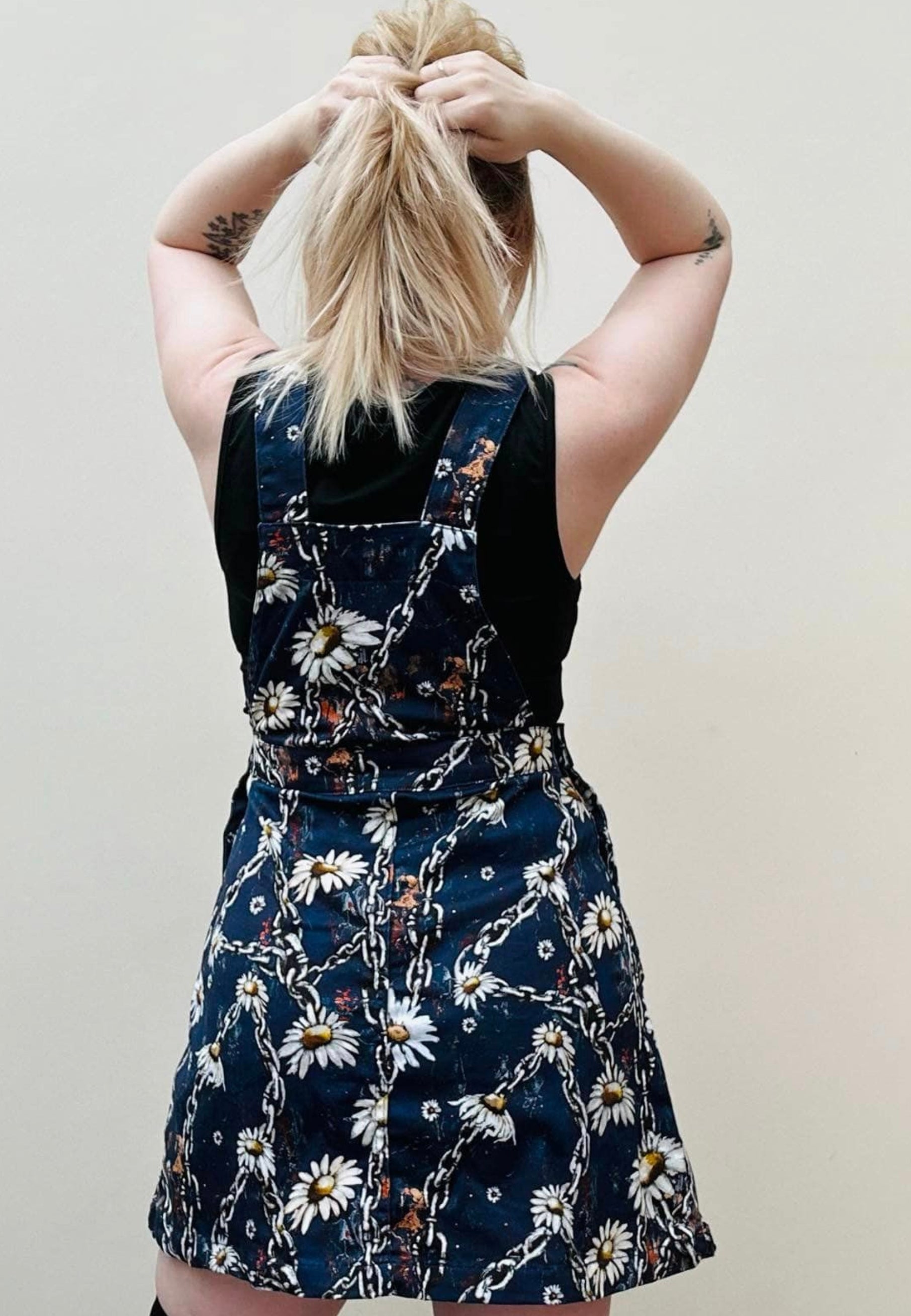 Back view of a woman wearing a deep blue pinafore dress with a unique urban grunge Daisy Chains print, featuring deep hip pockets and side button fastenings. View shows the shape of the pinafore from the back. Alternative styling.