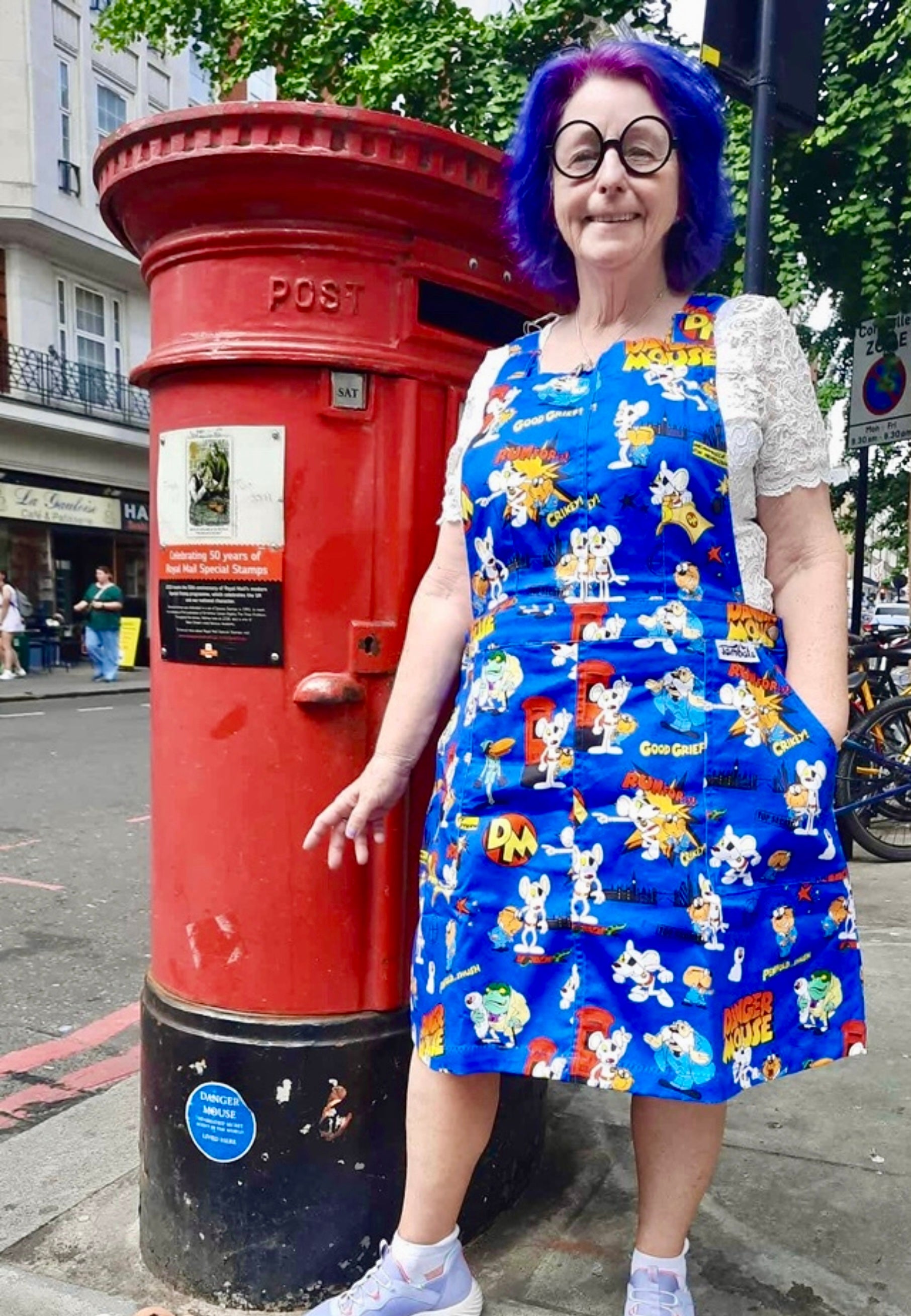 Woman wearing a blue pinafore dress with the 1980s cartoon characters from DangerMouse standing next to the Danger Mouse red postbox on a street in London. Model is wearing Penfold style glasses. 