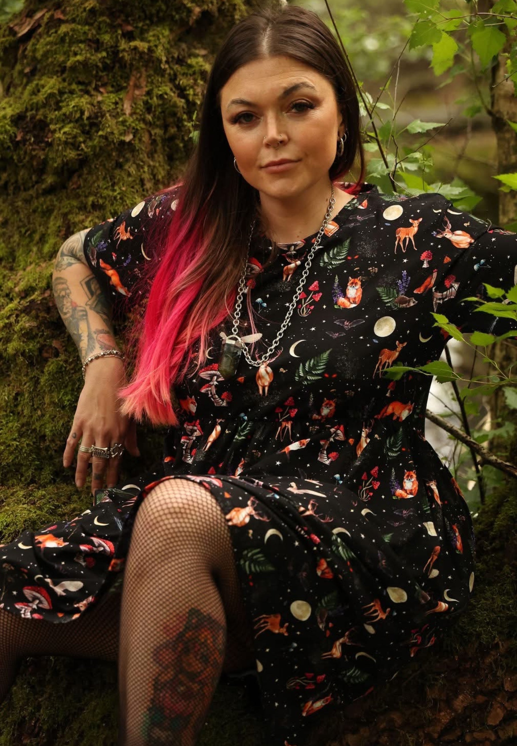 A woman with long dark hair and pink ombre ends sits outdoors against a mossy tree trunk, wearing a black dress with a colorful woodland print of foxes, deer, moons, and stars, along with fishnet stockings.