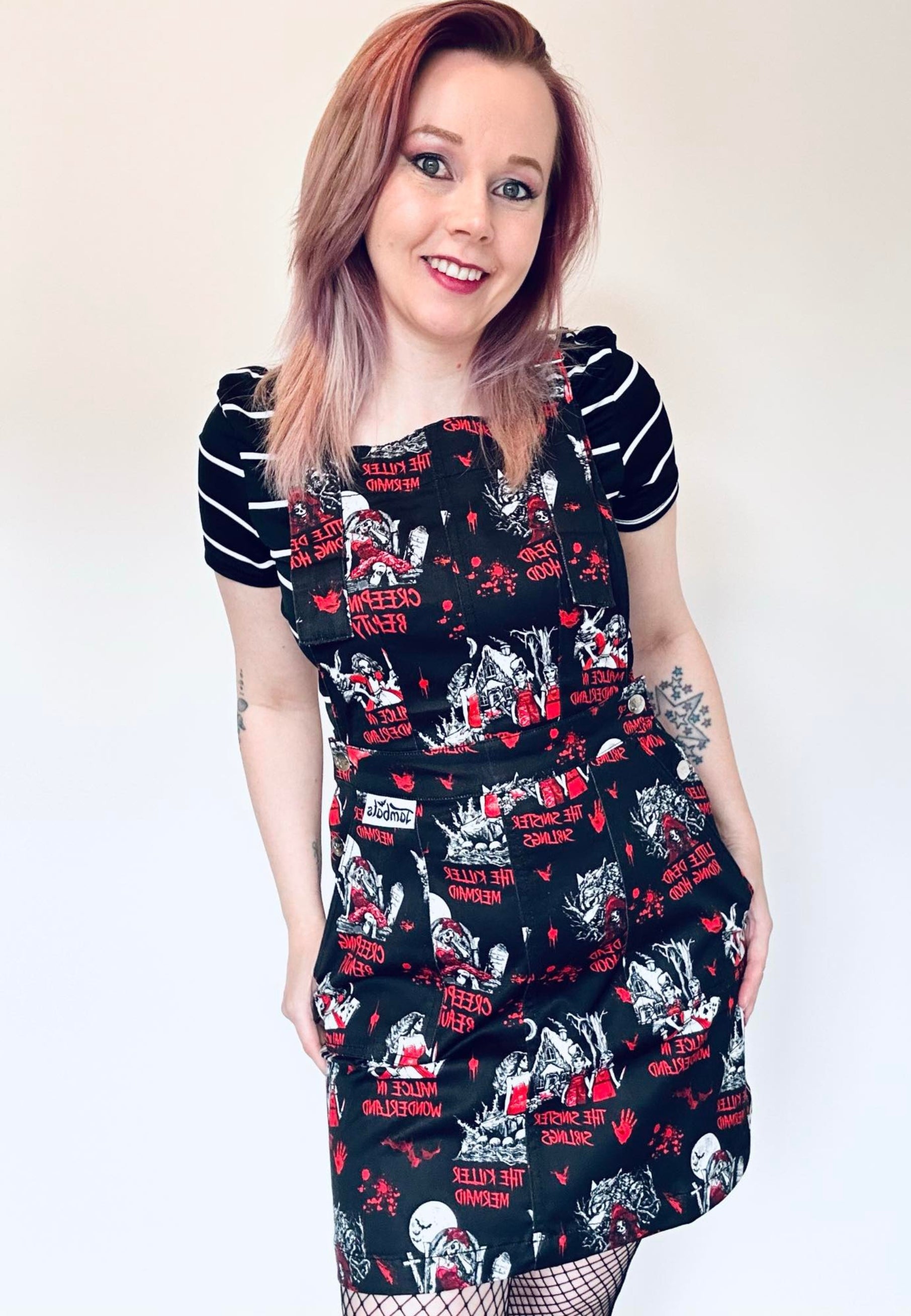 A woman wearing a black pinafore dress with a red and white character-based print, paired with a striped top underneath. Print on the pinafore is Jambats Twisted Tales which is a goth, horror print of dark fairytales. 