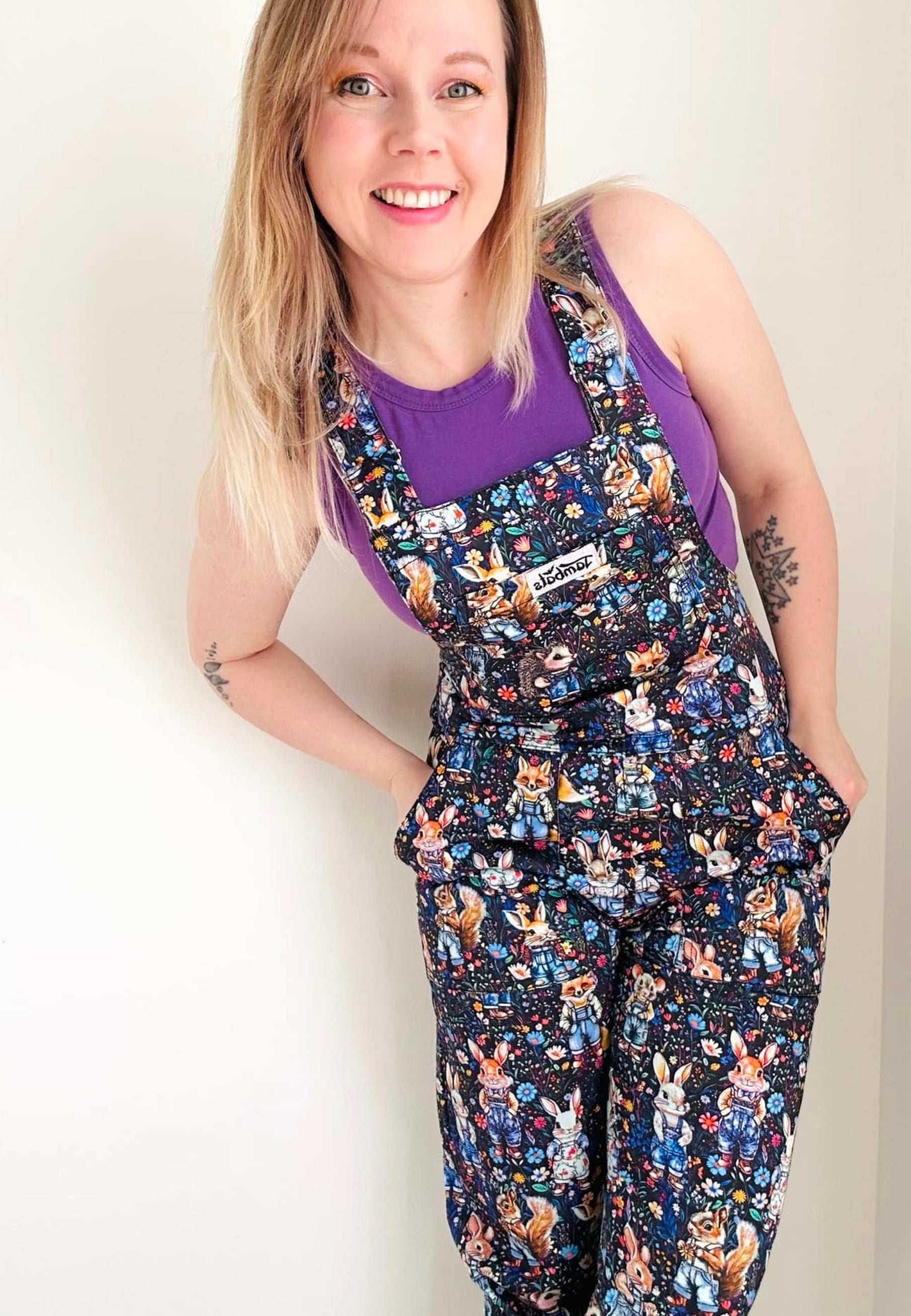 Woman wearing a pair of multicoloured cotton dungarees with a unique print of whimsical woodland creatures like a rabbit, fox, hedgehog, mole..all wearing denim dungarees on a floral background. Paired with a denim jacket. 