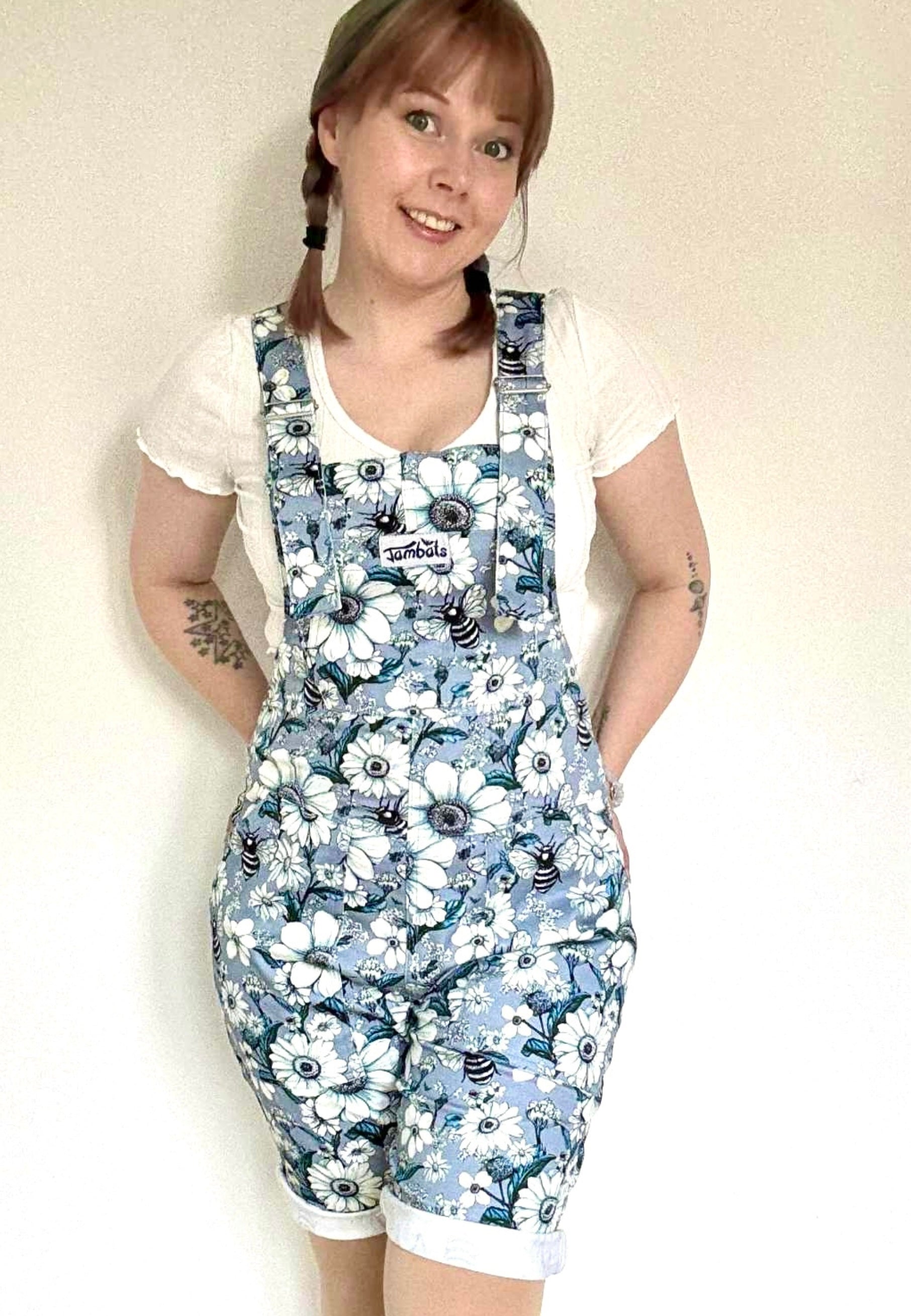 Woman wearing a floral daisy and bees patterned dungaree shorts against a plain background. JamBats dungaree shorts. 