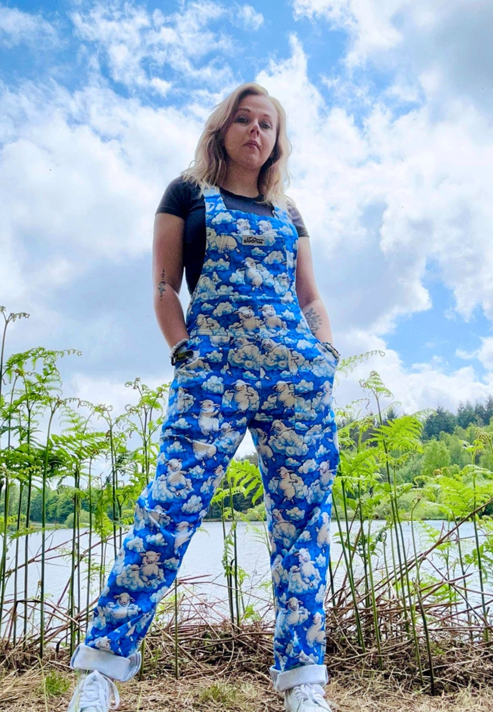 Model wearing a blue fluffy cloud and fluffy sheep patterned dungarees standing by a lake and trees with a blue sky and fluffy clouds behind them. 
