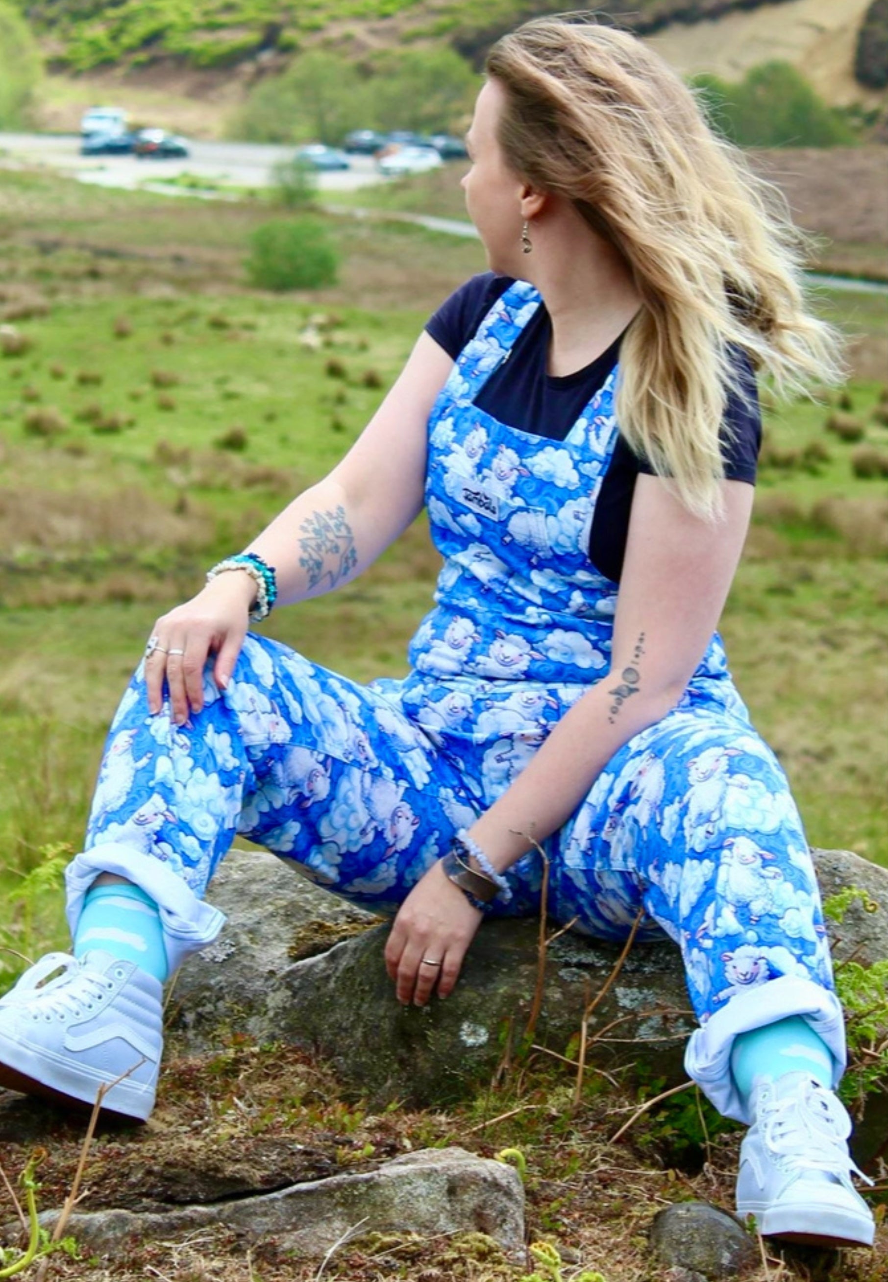 Model wearing a blue fluffy cloud and fluffy sheep patterned dungarees sitting on a rock in a grassy field.