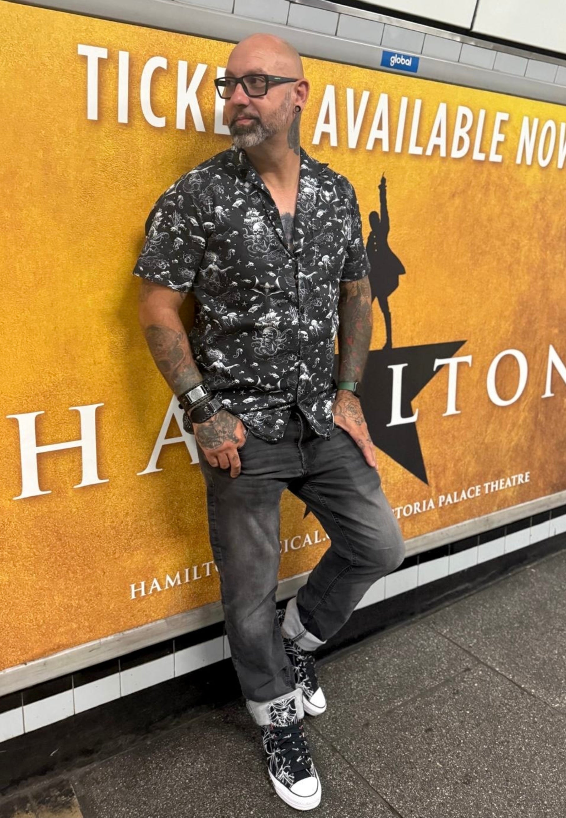 Man with tattoos leaning against a 'Hamilton' theater poster in a subway setting wearing Jambats black Abyss shirt and black jeans. 