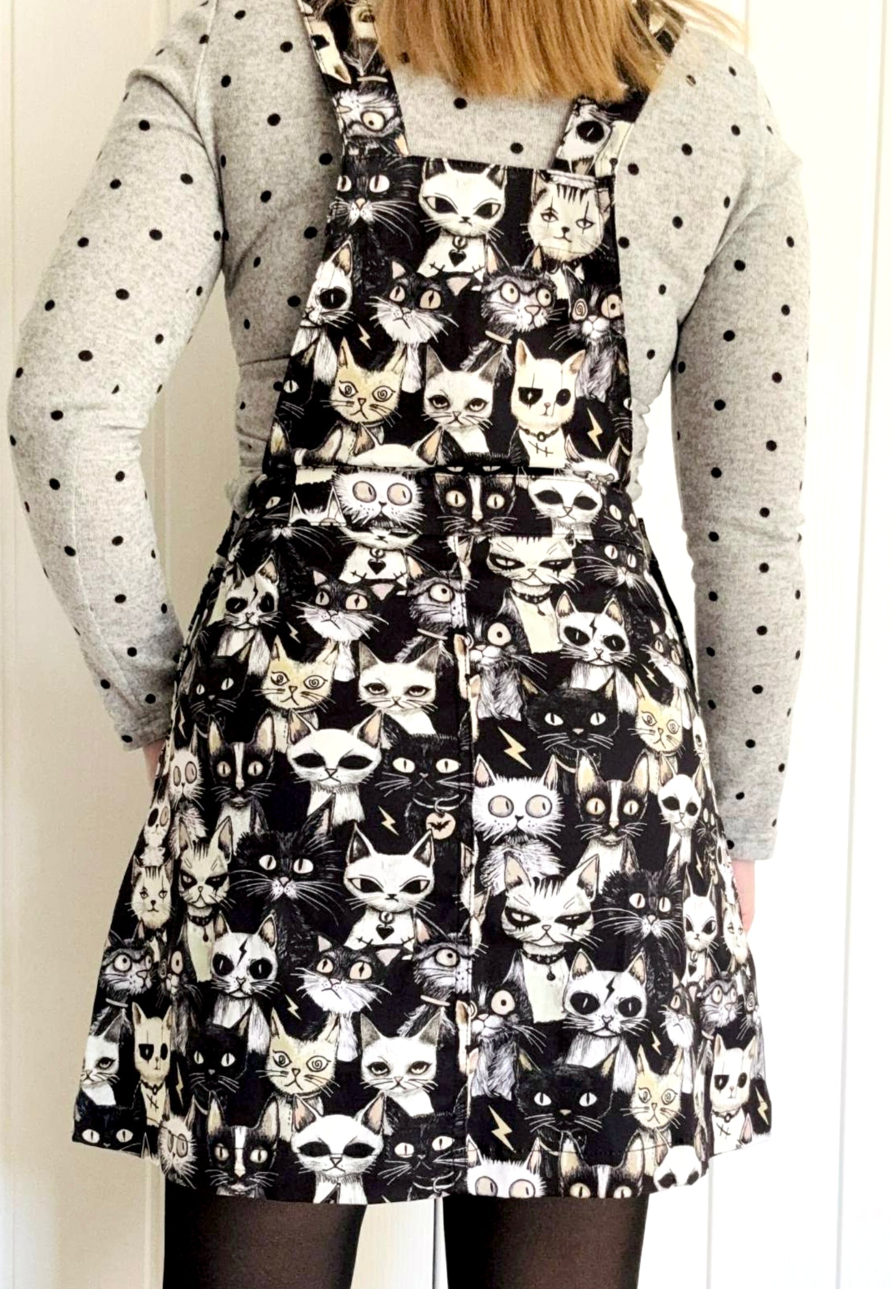 Back view of a model wearing a black and white pinafore dress with a print of cute but gothic alley cats. Alternative cats print. 
