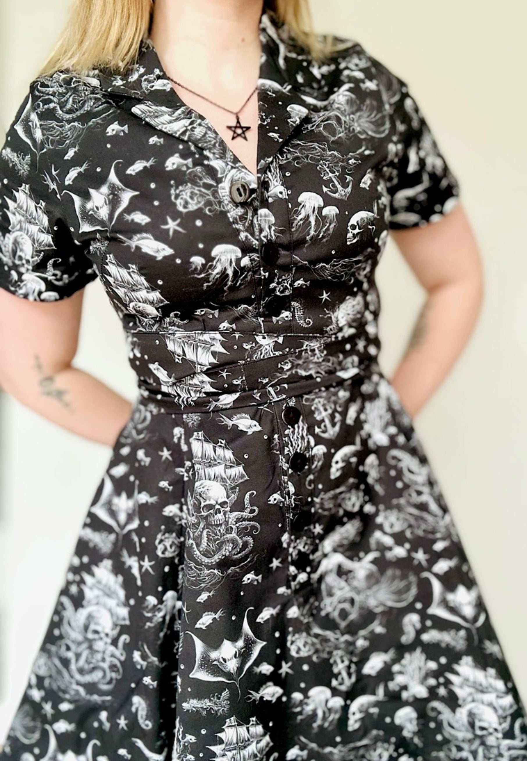 Woman wearing a black shirt dress with a beautiful gothic print of under the sea. Ghostly sirens and sunken skull pirate ships. Studio photo against a plain background. Goth style dress.