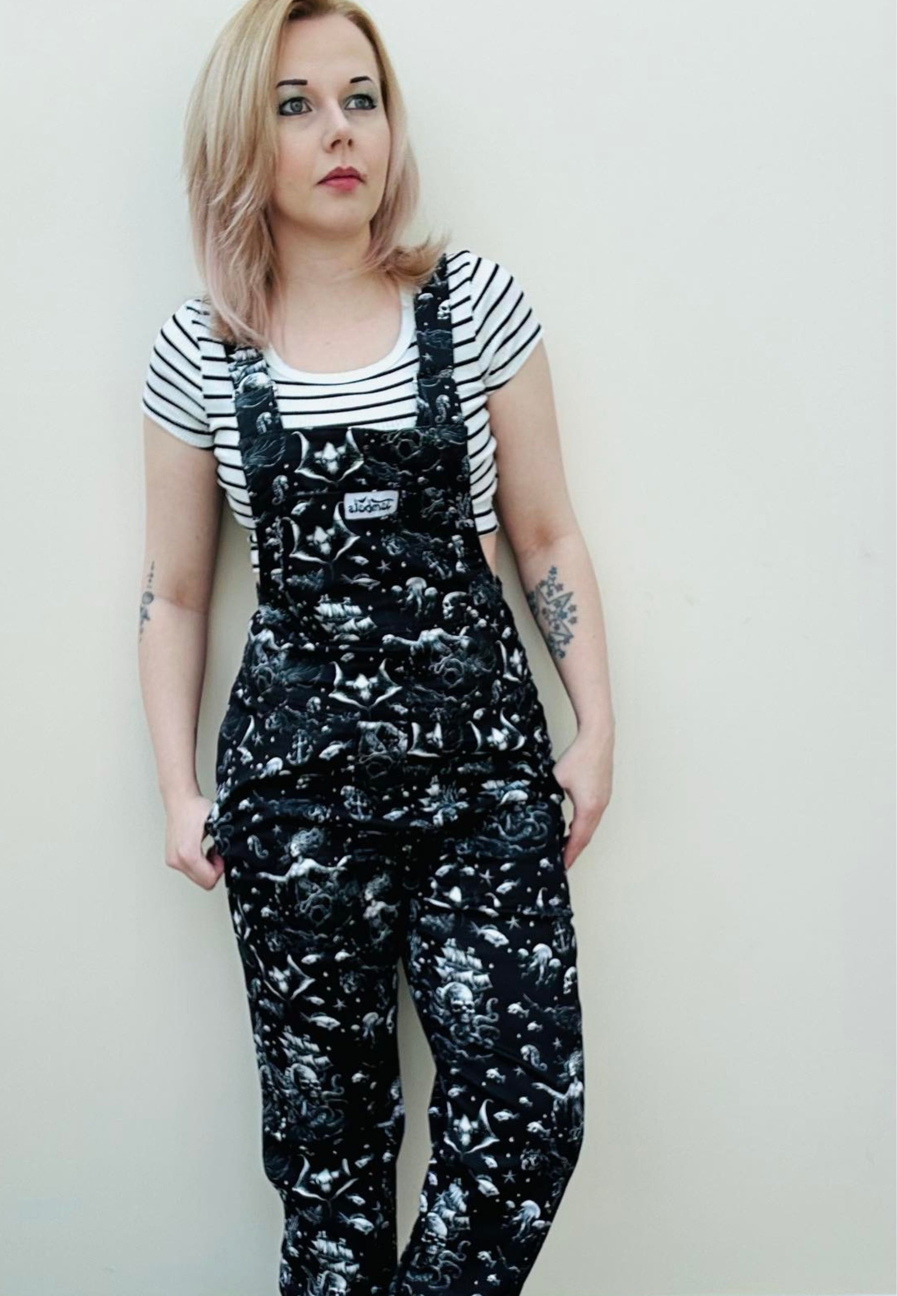 A woman wearing black dungarees with a pattern of ghostly pirate skull ships and eerie sirens. Styled with a black and white striped top 