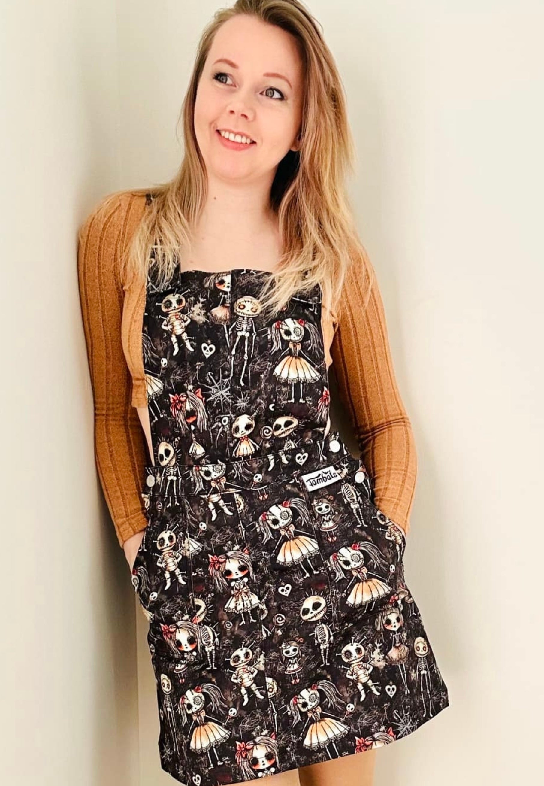 A woman wearing a stretch cotton twill pinafore dress with a haunted dolls print, featuring deep hip pockets and side button fastenings with unique bat buttons. The dolls print has cute but creepy dolls in a dusty, spiders web attic setting. 