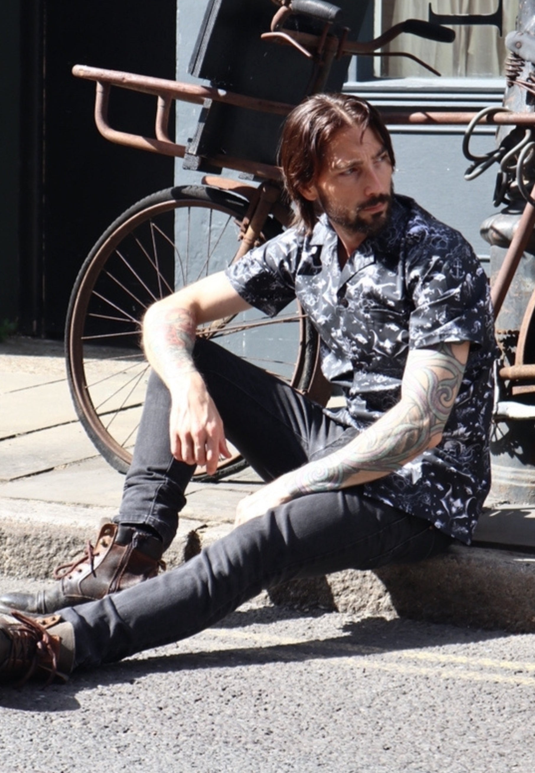 Man sitting on a curb with bicycles in the background. Man is wearing Jambats Into the abyss gothic under these shirt and black ripped dreams. 