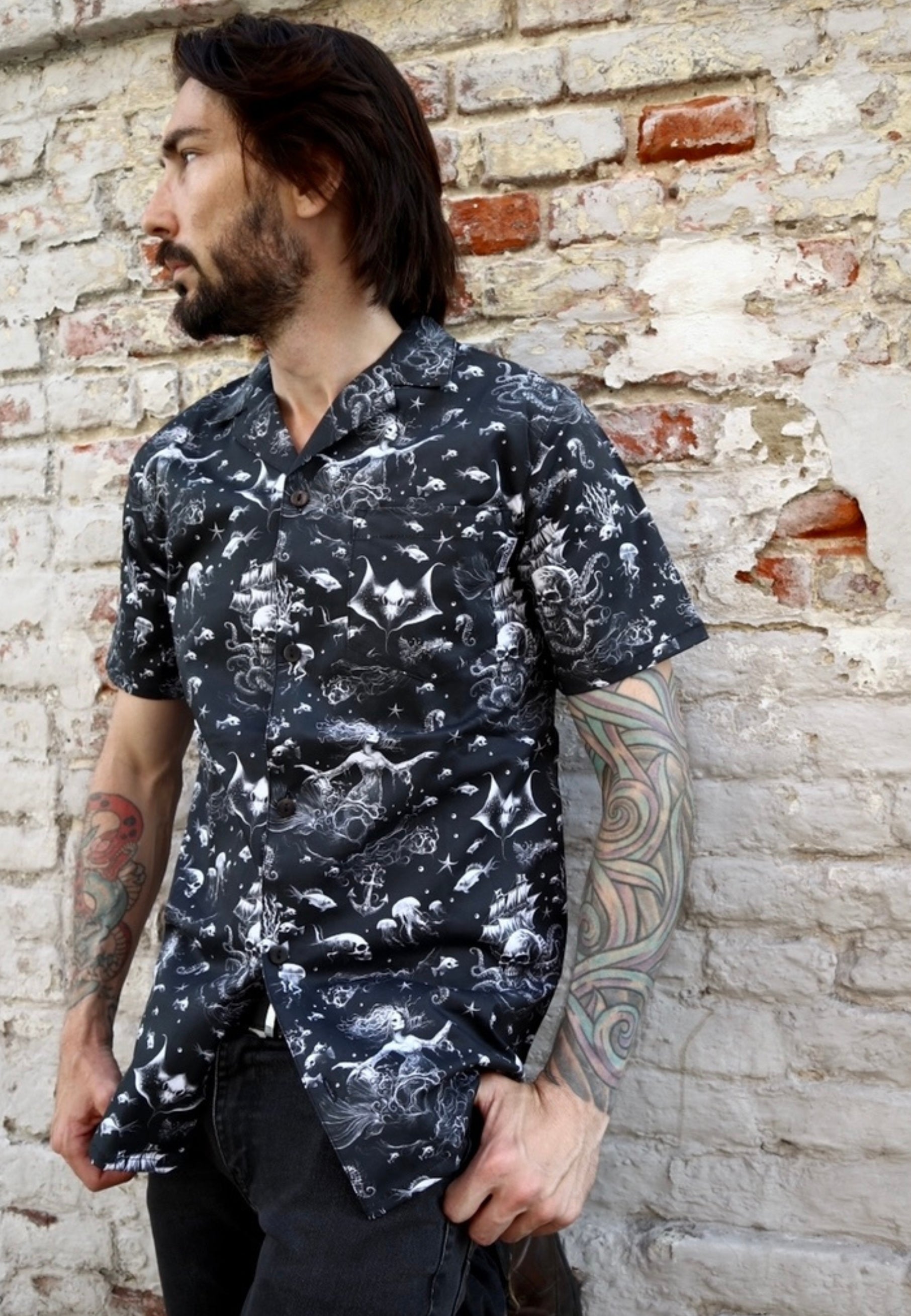 Man wearing a black shirt with a gothic under the sea print..ghostly mermaids and pirate ships. Man is standing in front of a whitewashed brick wall