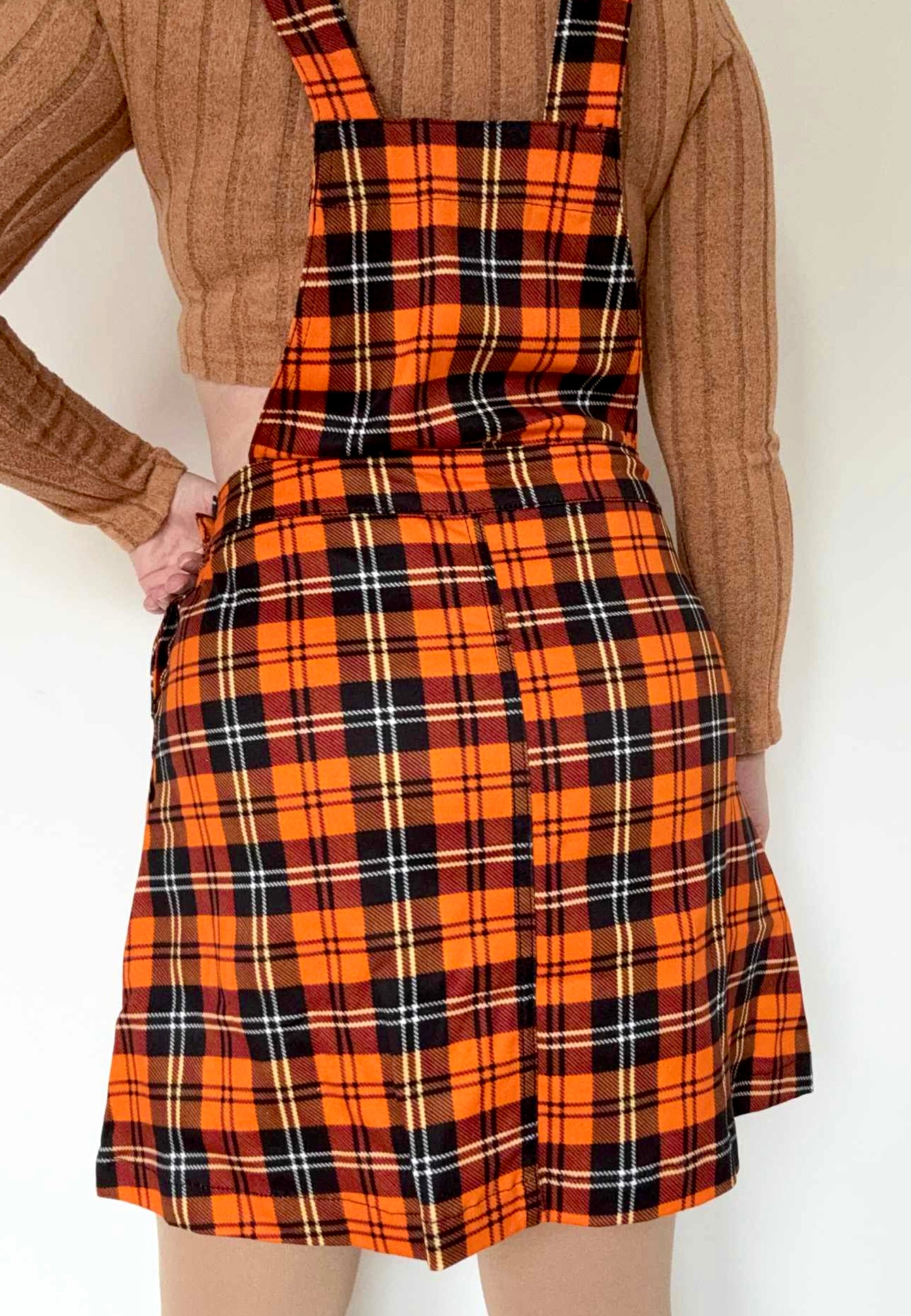 Back view of a woman wearing an orange tartan pinafore dress over a sweater against a white background.