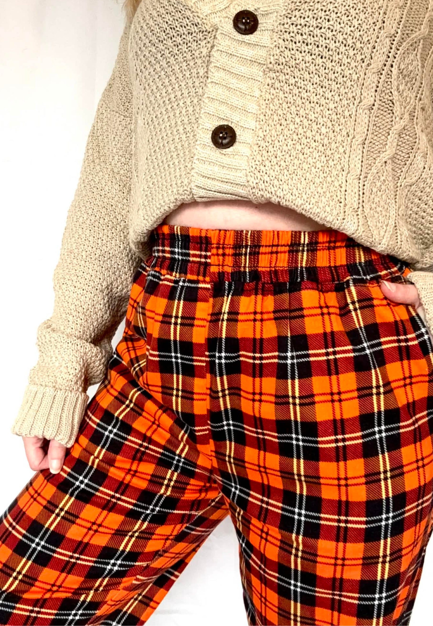 Person wearing a beige knitted slouchy cardigan and orange tartan trousers. Close up of the trousers on a white background