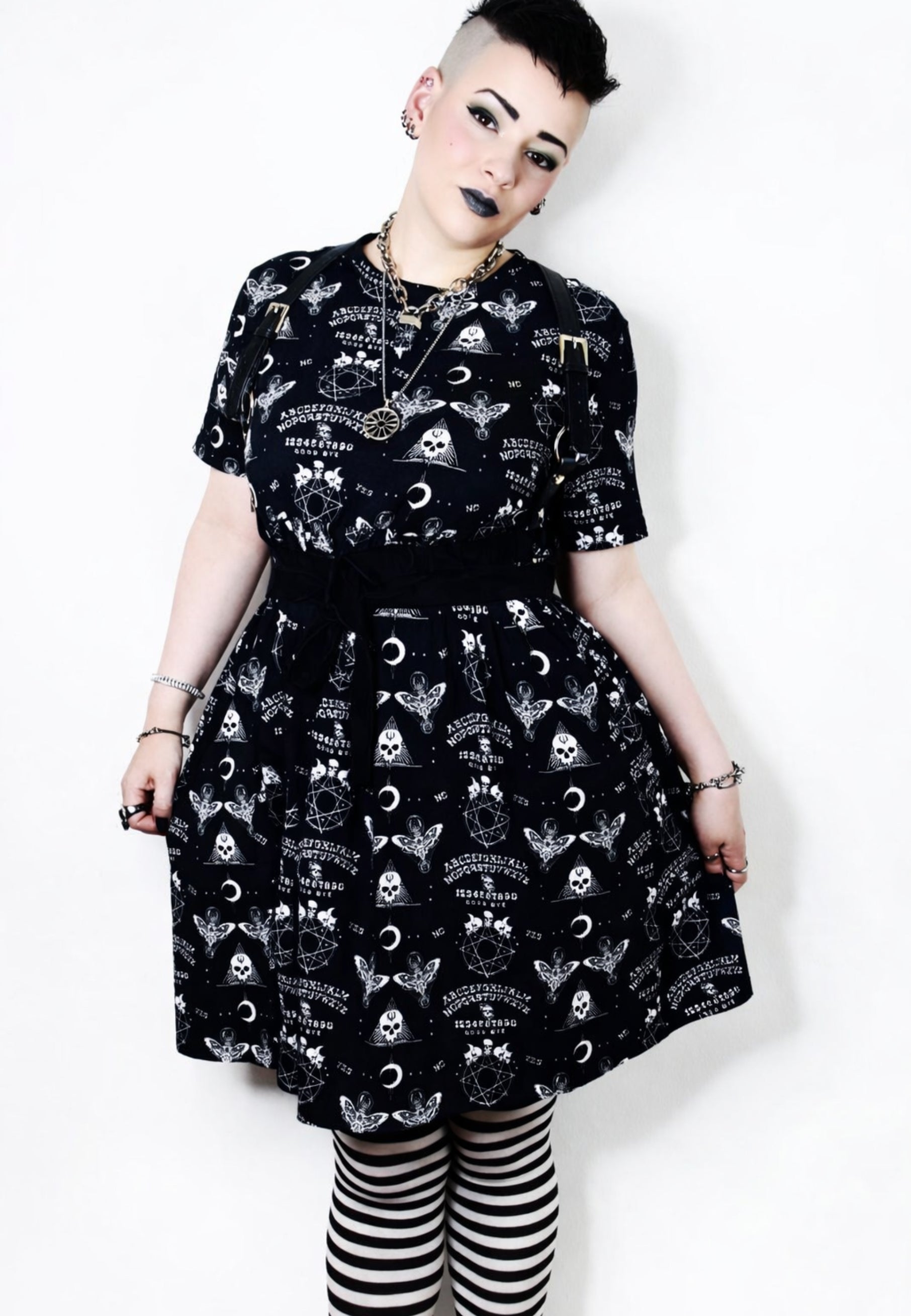 Woman wearing a black smock dress with gothic Ouija print pattern against a plain background. Studio photo. Model is also wearing black and white striped tights. 