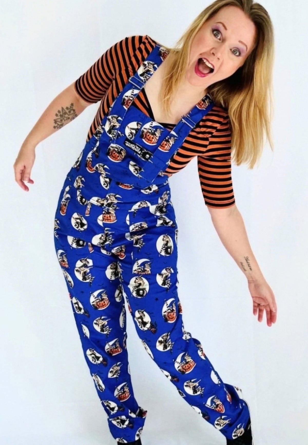 A woman modelling a pair of blue dungarees covered in a repeat print of retro spooky pin up witches, worn over a black and orange striped t shirt against a white studio background.