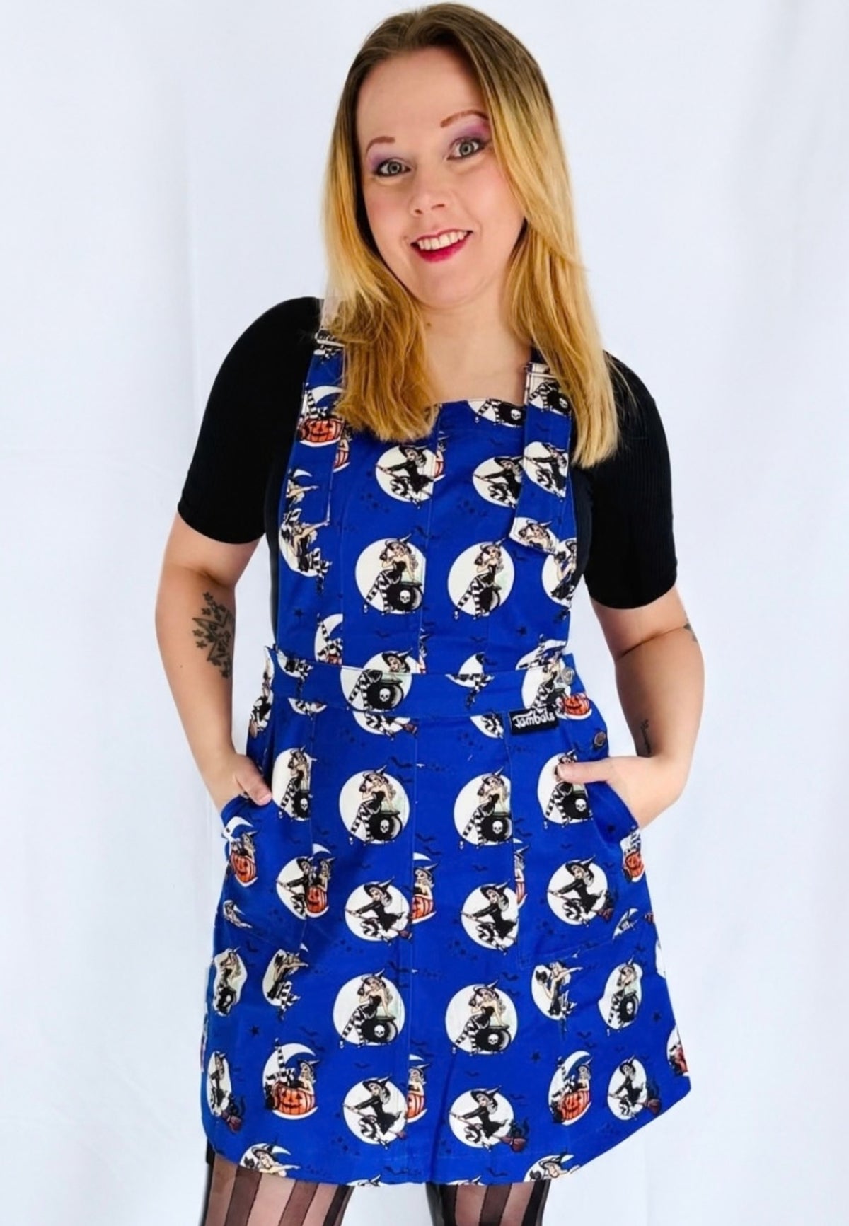 A woman modelling a blue pinafore dress with retro glam print if pin up witches against full moons. Model is wearing black striped tights and standing against a white background 
