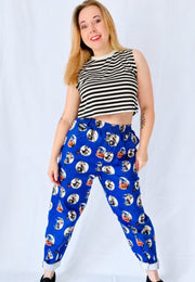 A woman modelling a pair of blue trousers with an all over pattern of spooky retro pin up witches, paired with a black and white striped crop top, against a white studio background.