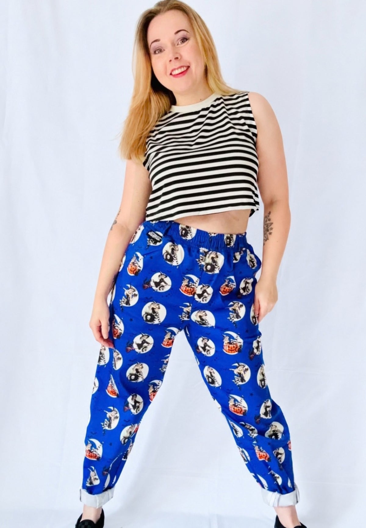 A woman modelling a pair of blue trousers with an all over pattern of spooky retro pin up witches, paired with a black and white striped crop top, against a white studio background.