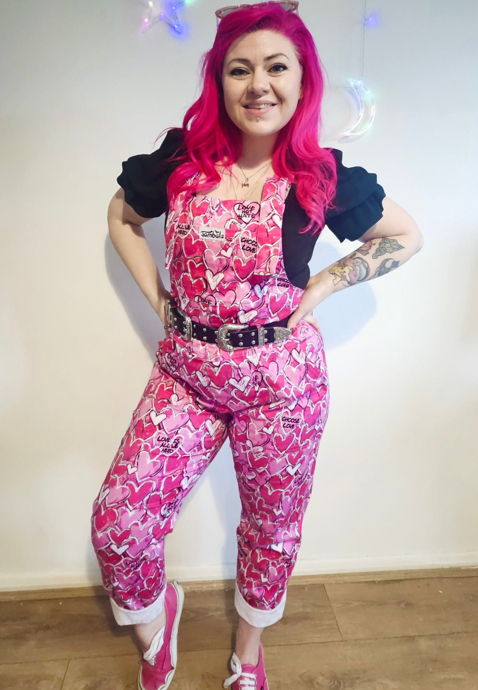 A woman with beautiful dark pink hair wearing a pink and white heart patterned dungarees with a black top, standing against a white background. 