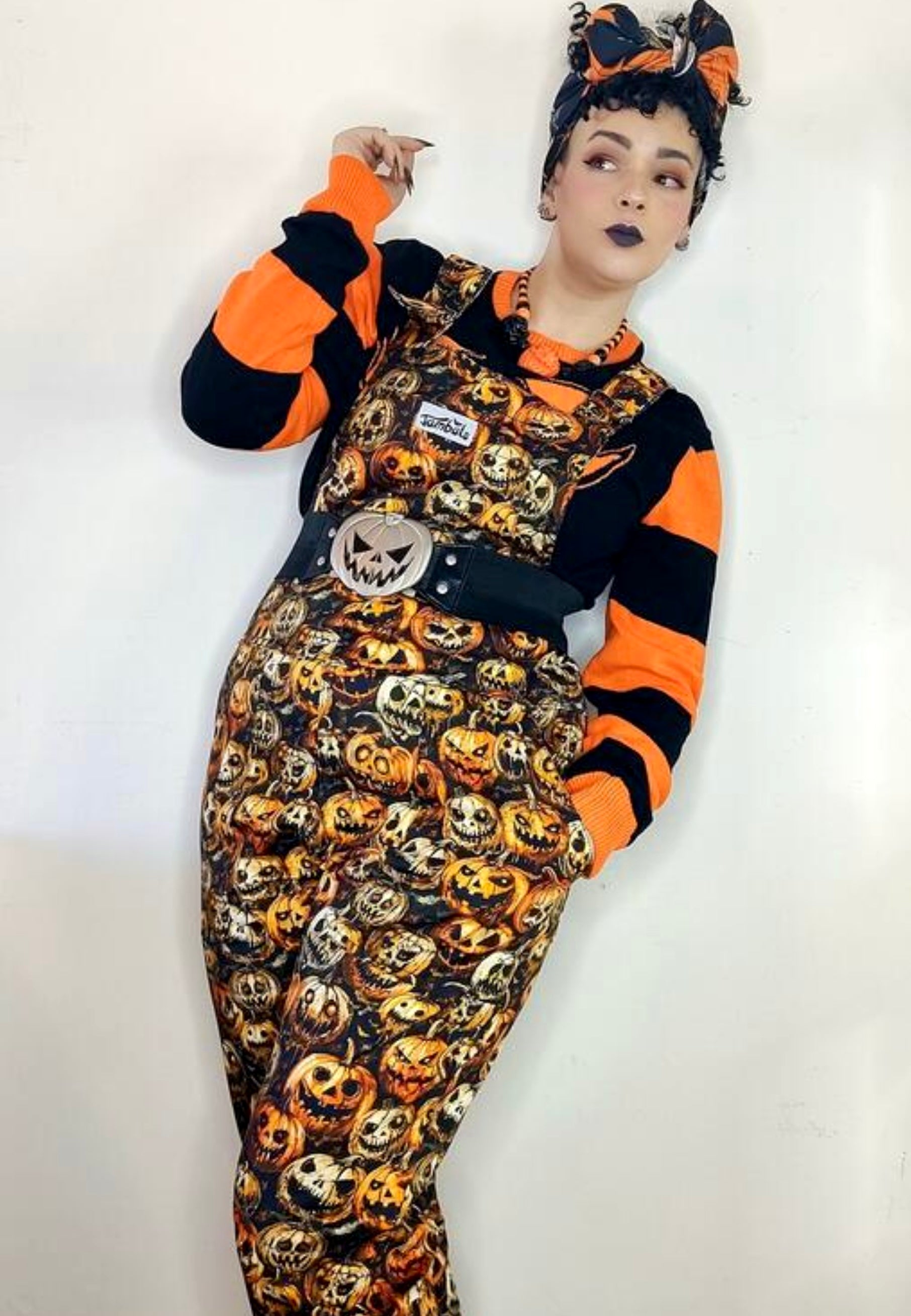 Model wearing Halloween themed dungarees overalls with illustrated creepy pumpkins print. Model is also wearing orange and black striped jumper and hair tie. Quriky retro spooky Hallween style stduio photo on white background.