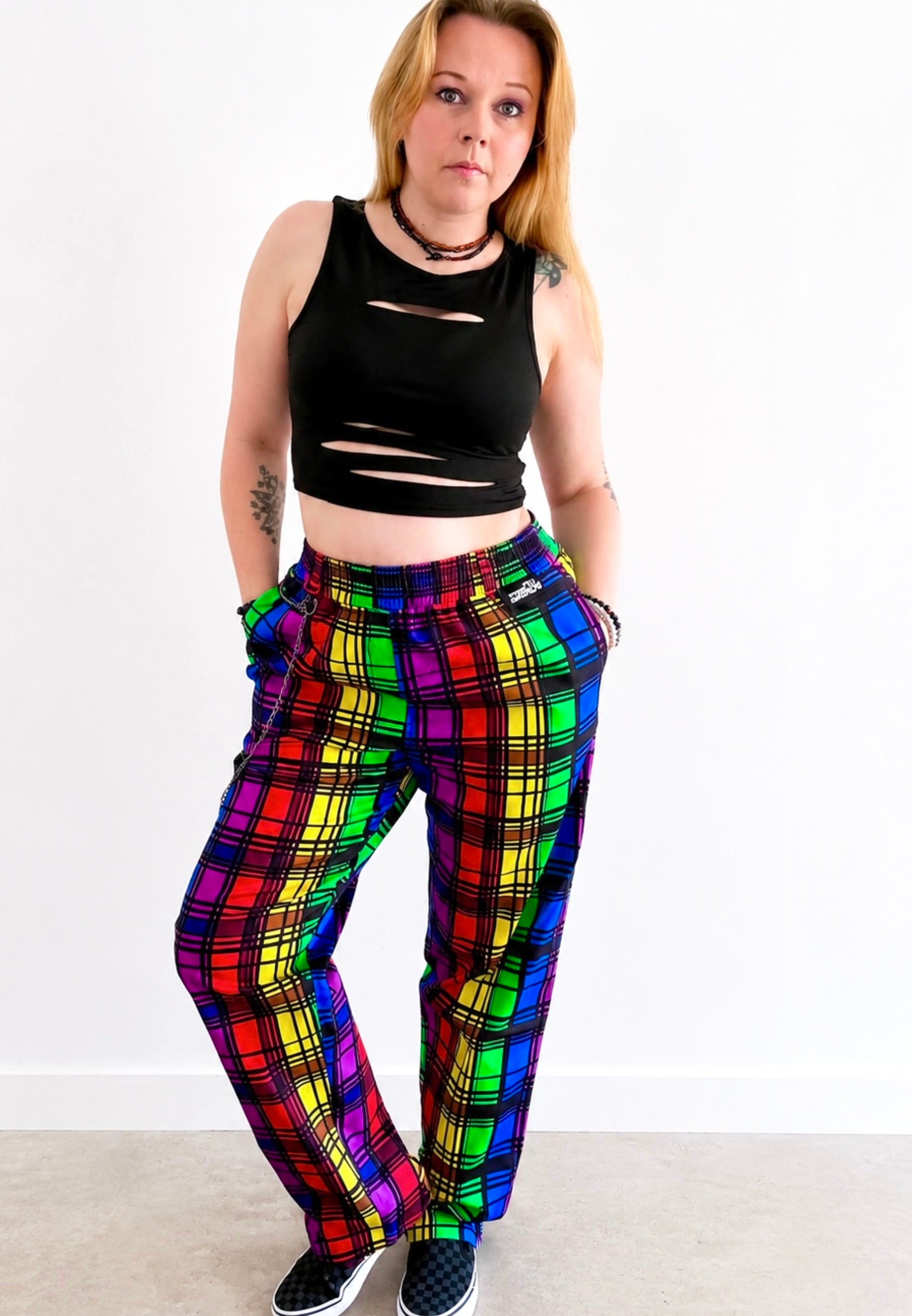 Person wearing a black ripped crop top and vibrant rainbow tartan trousers with a punky chain on belt looks. Photo is on a plain background