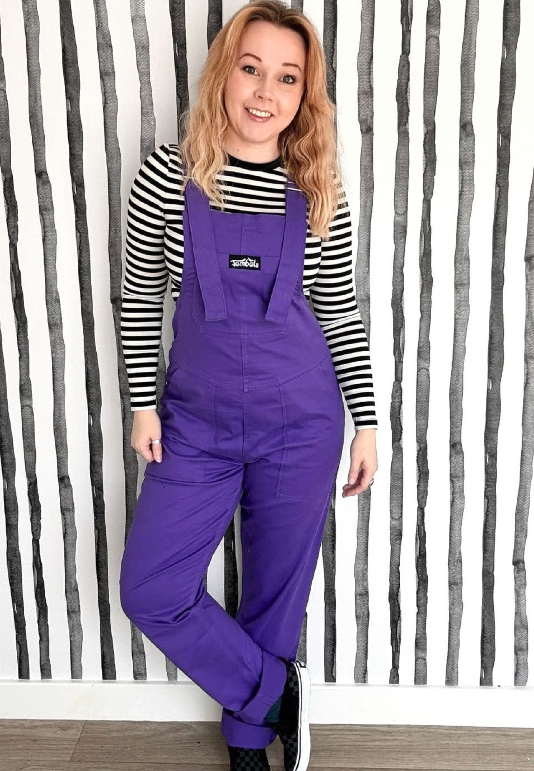 Woman wearing purple dungarees with a striped shirt against a striped wall background
