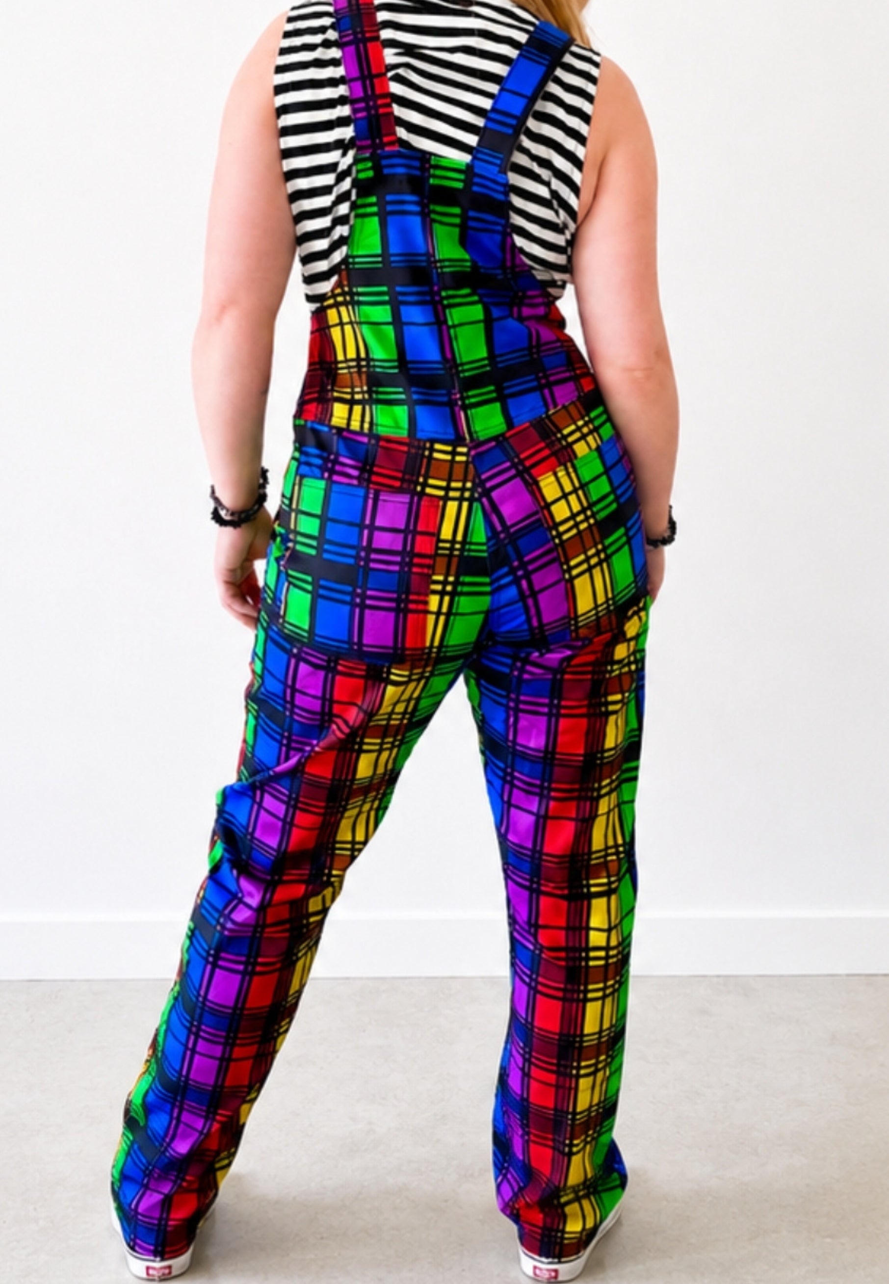 Woman wearing bright coloured rainbow tartan dungarees with a black and white striped top. Showing dungarees from the back. Woman is standing against a white background