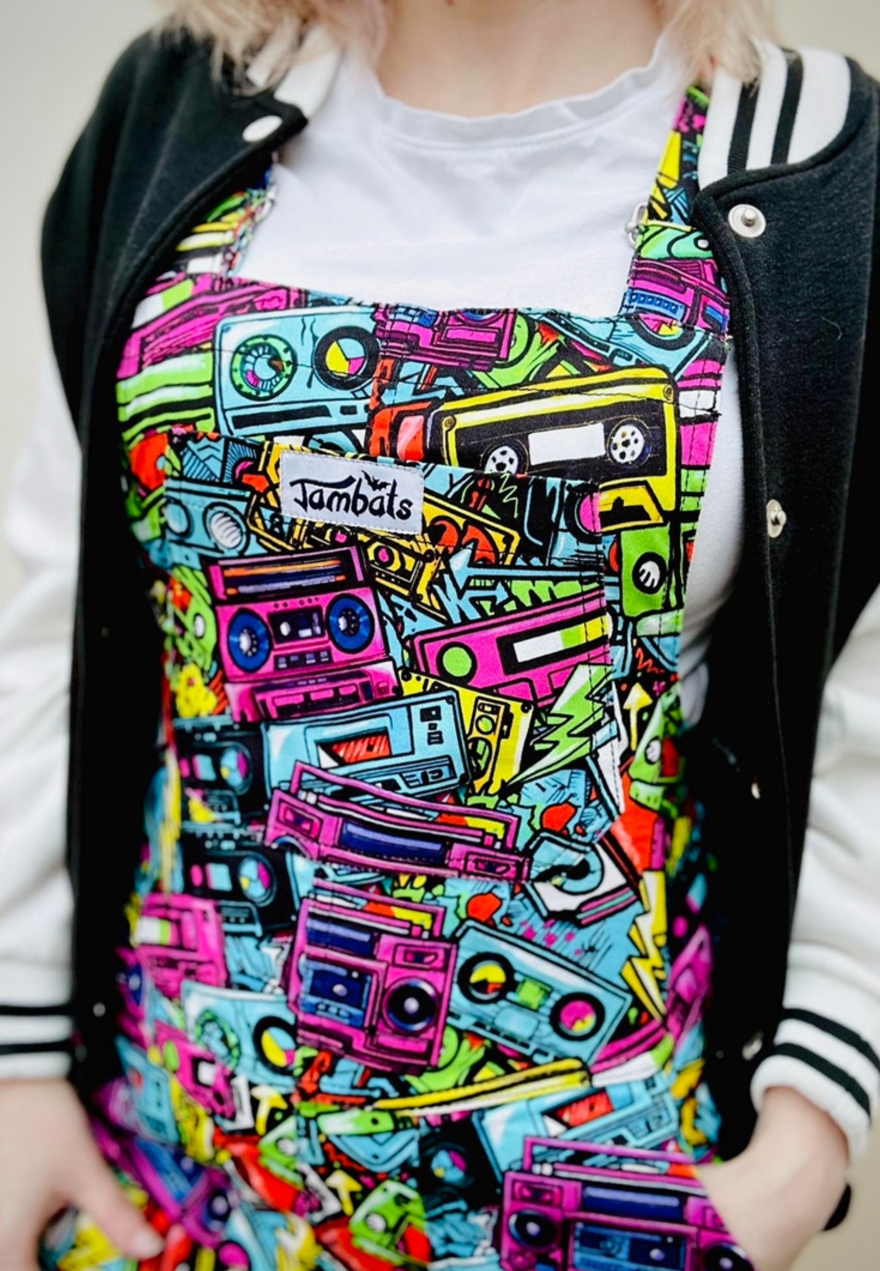 Woman wearing bright coloured dungarees with a retro 80s 90s print of graffiti street art music cassettes and ghetto blasters. Casual styling. Close up image of the dungaree print. 