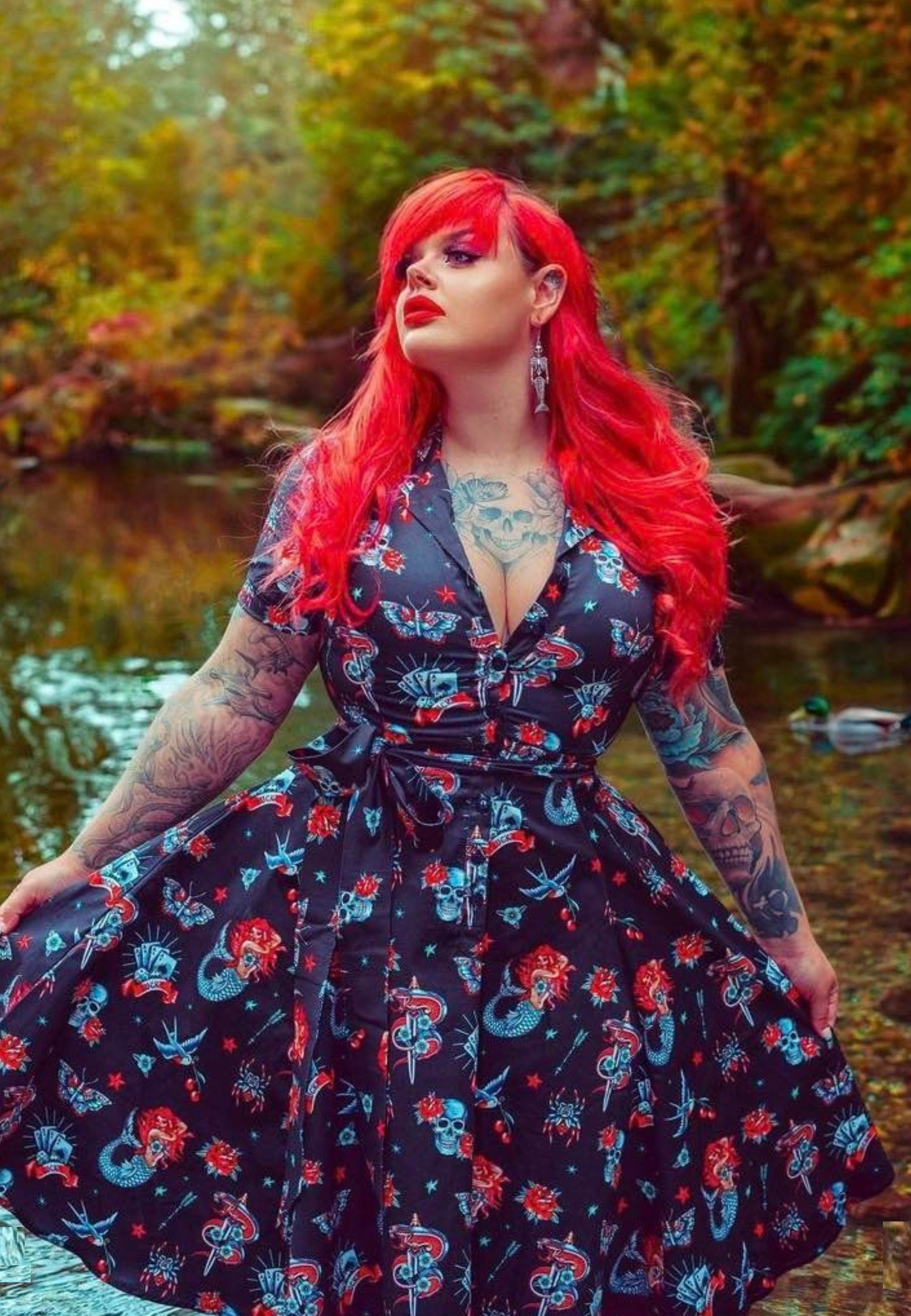 A woman with red hair wearing a retro shirt dress with a traditional tattoo print in shades of red, teal, and blue. The dress features a full skirt with button shirt bodice, hidden hip pockets, and a fabric belt.