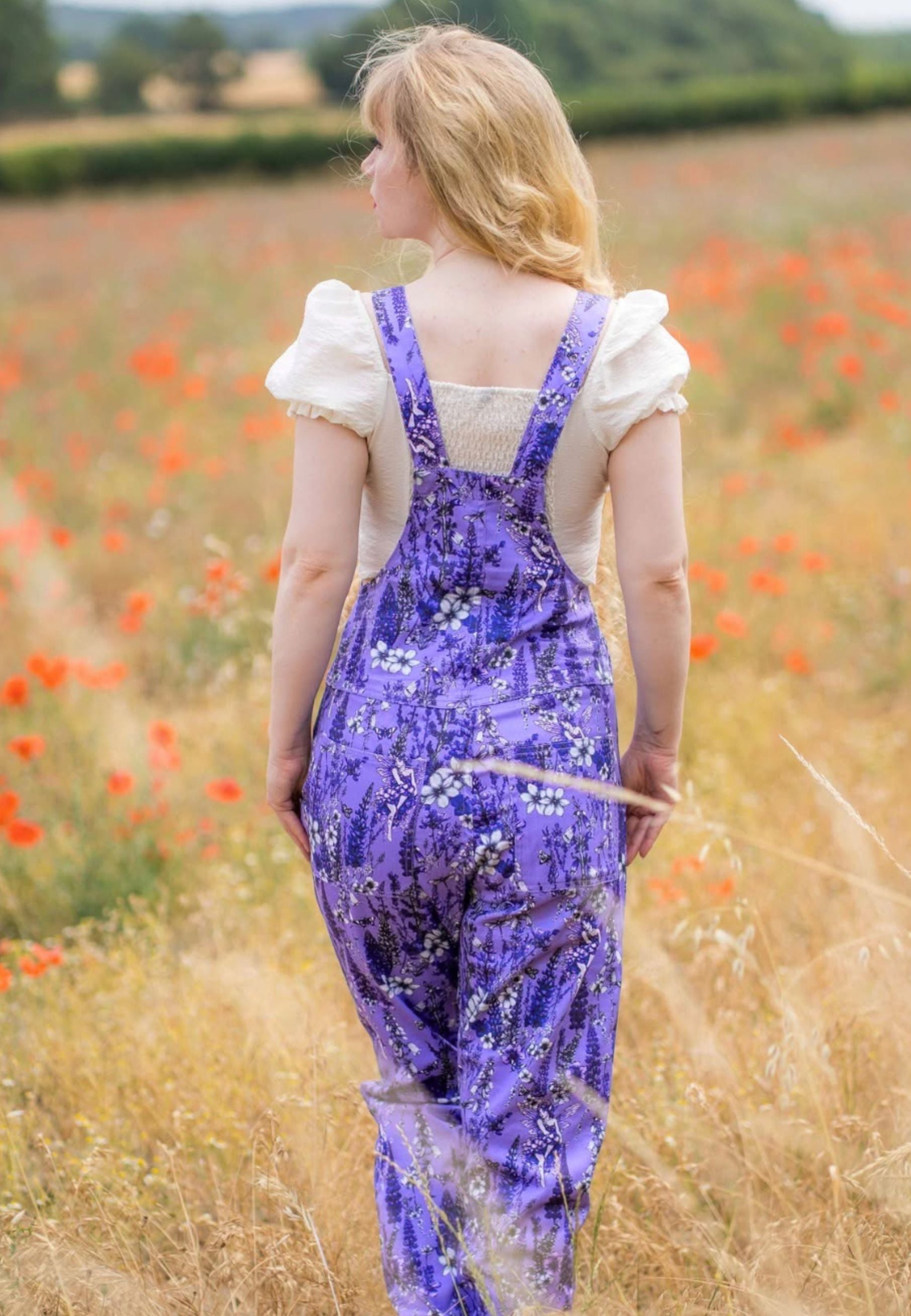 A woman standing in a field of corn, wearing a lilac overall dungarees with a white ruffled top. The dungarees have a beautiful whimsical print of wild flowers and flower fae. Photo shows model and dungarees from the back 