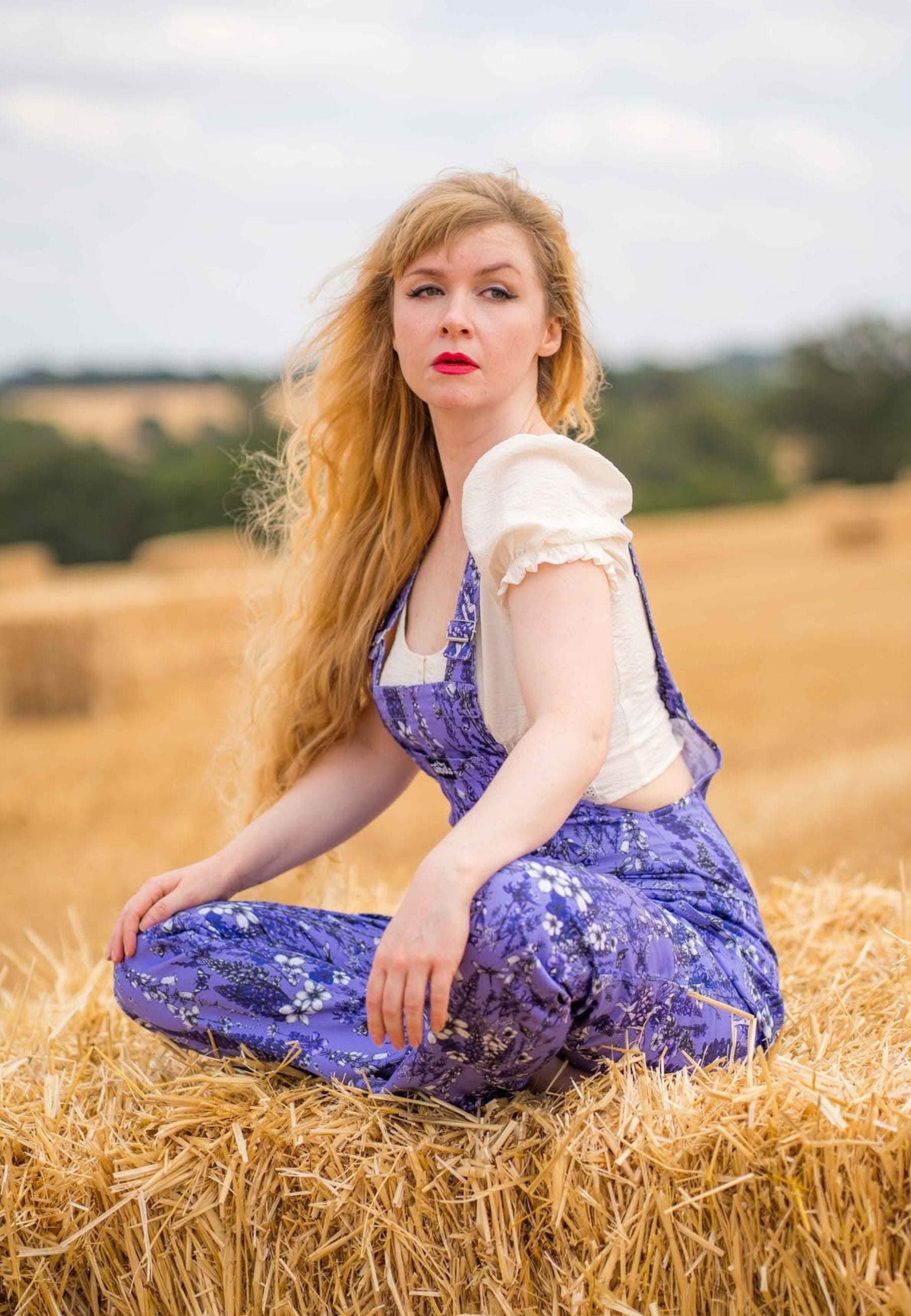 A woman with long blonde hair sits cross legged on a hay bale in a field, wearing purple floral overalls dungarees over a cream puff-sleeve top.