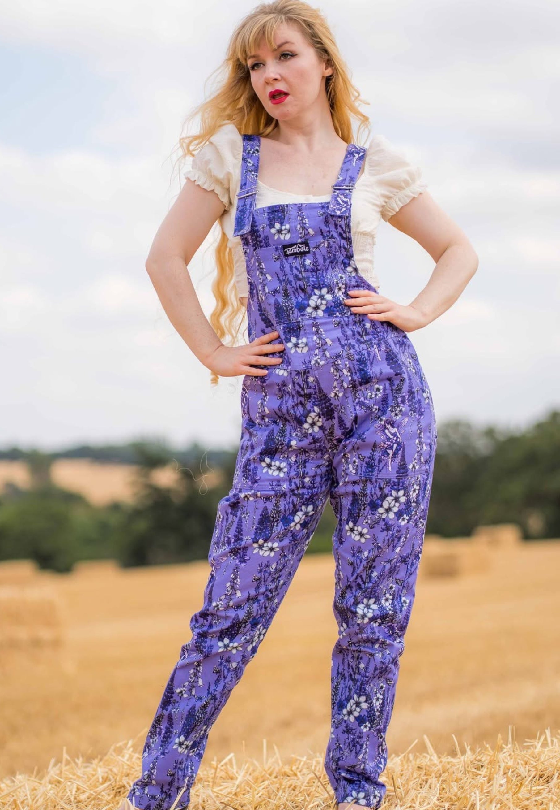 A woman stands outdoors in a field wearing purple floral overalls over a cream short-sleeve top. The overalls have a purple base with a whimsical floral pattern which has flower fae in it. 