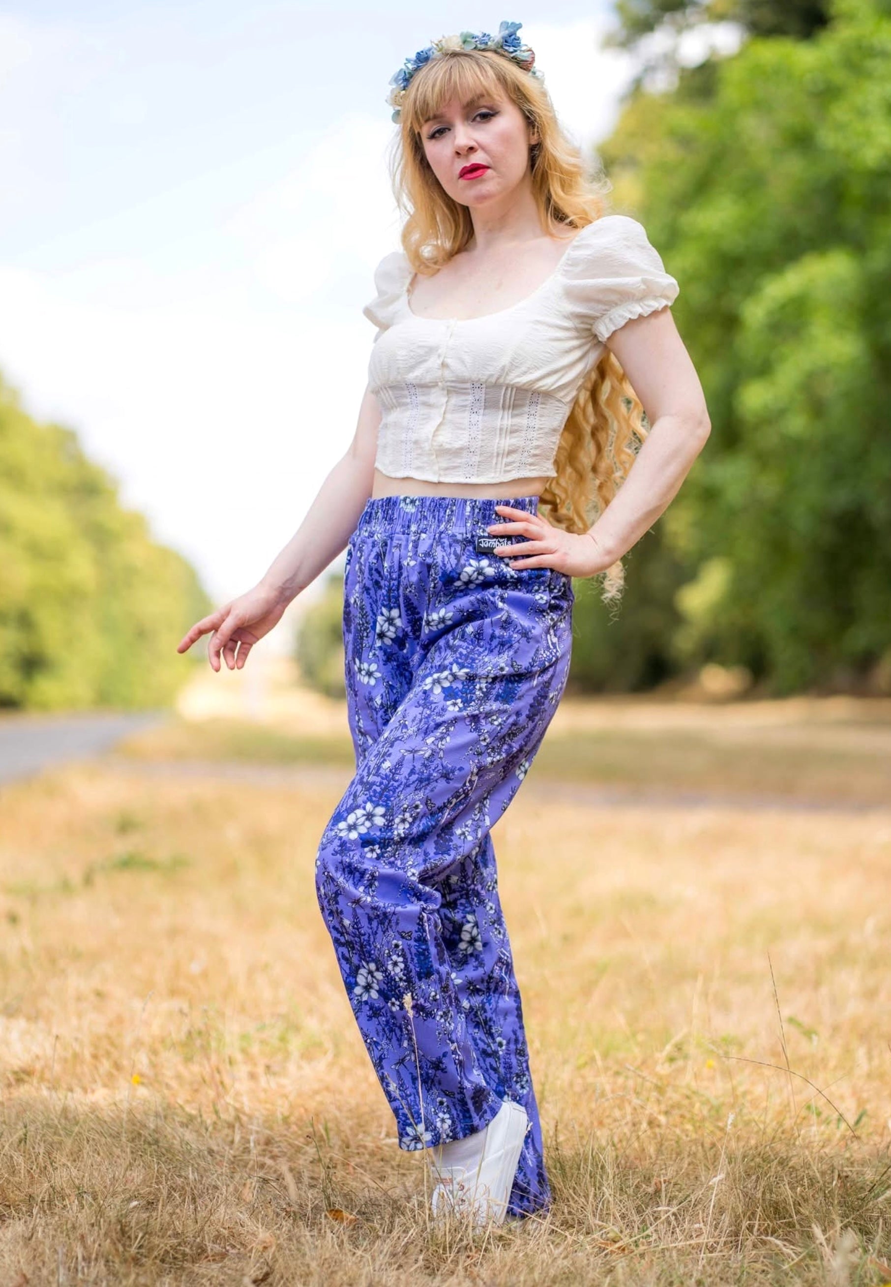 A woman standing outdoors in a grassy field wearing a cream puffsleeve crop top, purple wild flower fae print trousers and a blue flower crown.