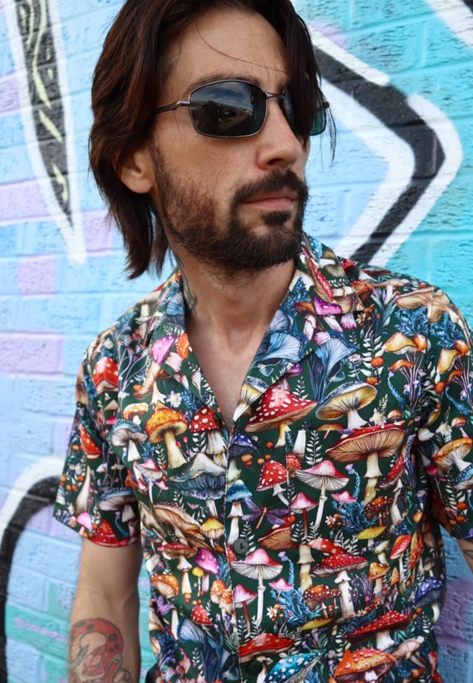 Man wearing a colorful mushroom-patterned shirt in front of a graffiti-covered wall.