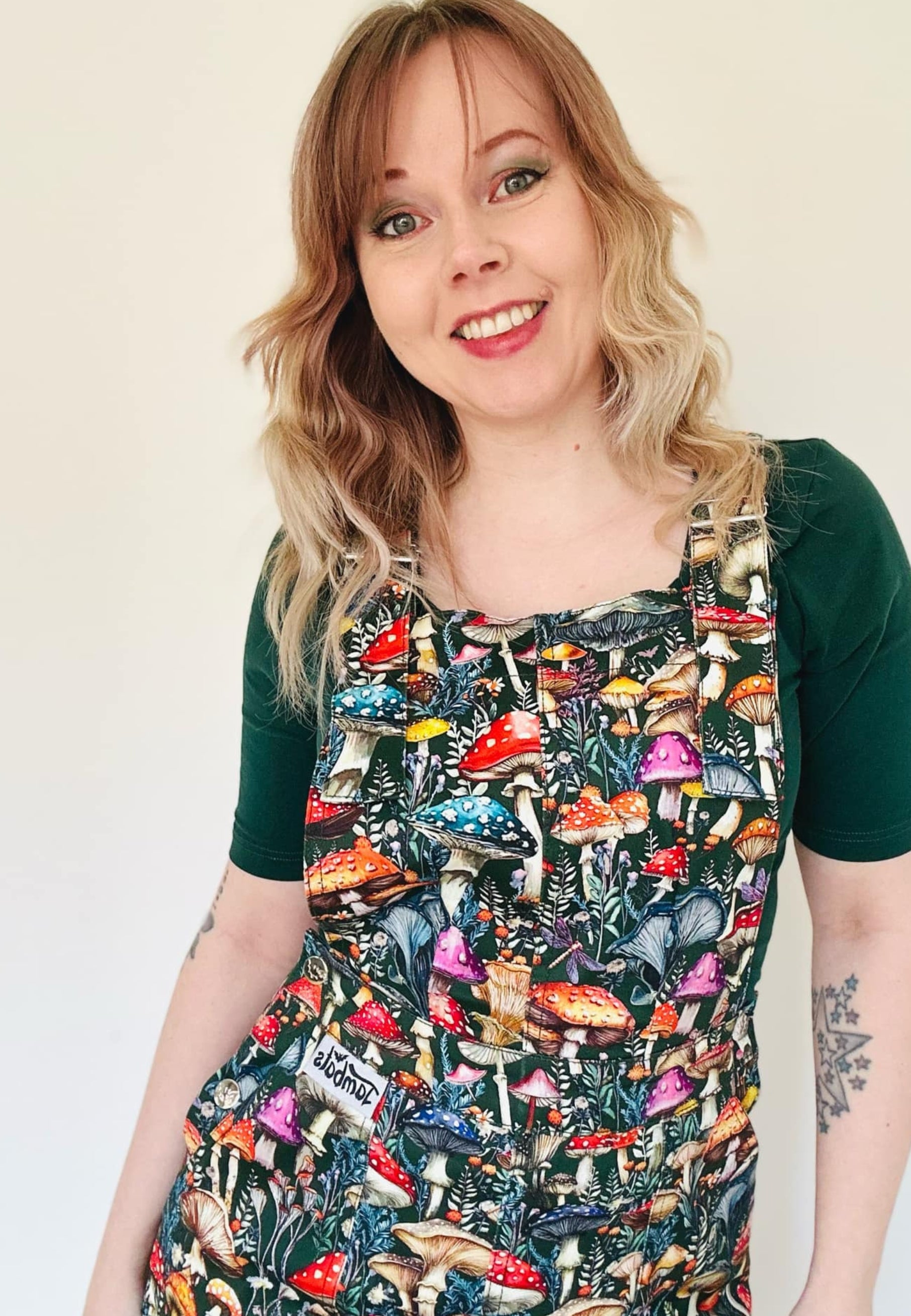 A person wearing a dark green pinafore dress with a vibrant mushroom print with mushrooms and toadstools. Studio photo on a plain white background. 