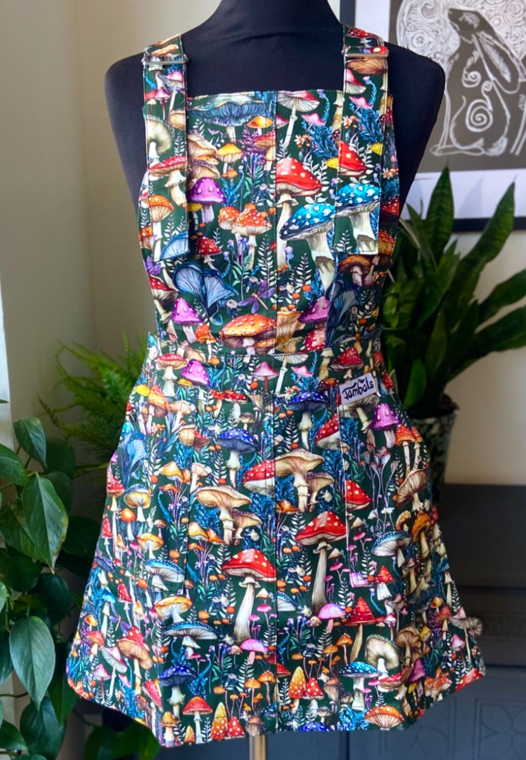 A mannequin with a dark green pinafore dress with a vibrant mushroom print with mushrooms and toadstools. Whimsical hare nd moon print picture in the background and houseplants. 