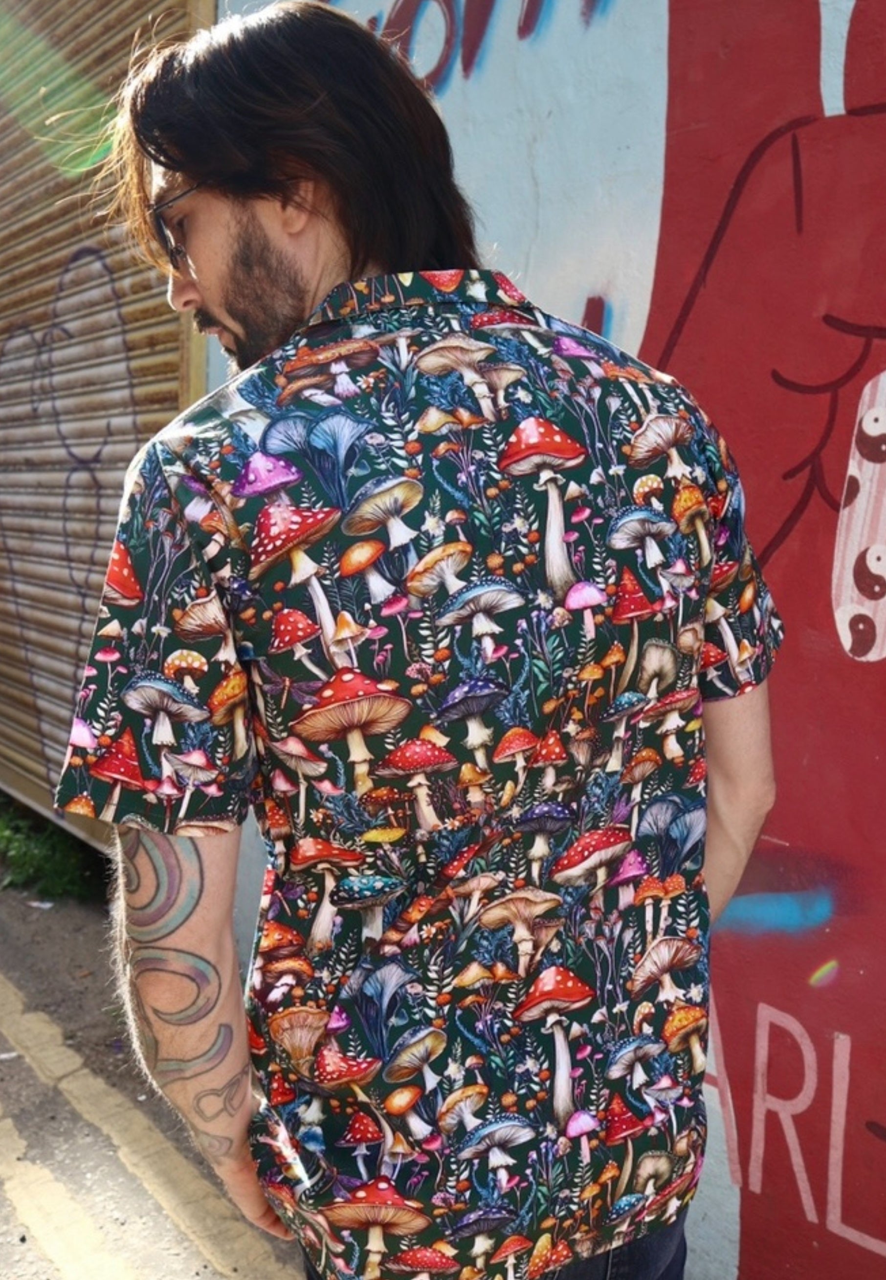 Back view of a man wearing a colorful patterned mushroom print shirt and black jeans standing in front of a graffiti-covered wall.
