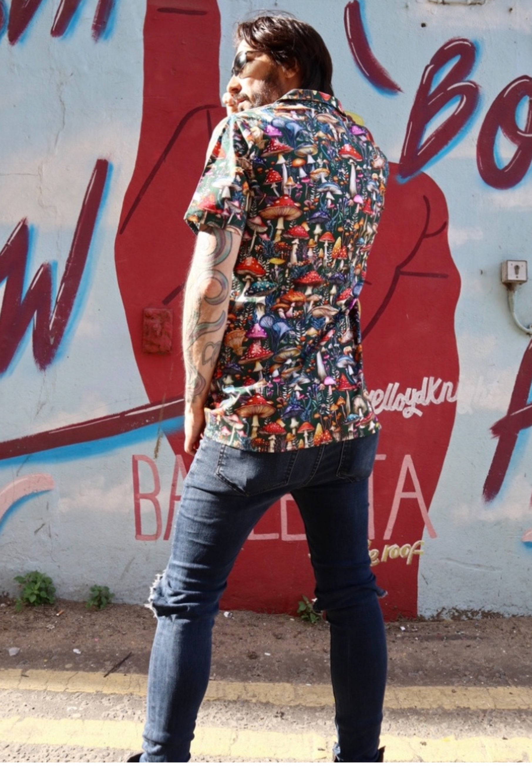 Back view of a man wearing a colorful patterned mushroom print shirt and black jeans standing in front of a graffiti-covered wall.