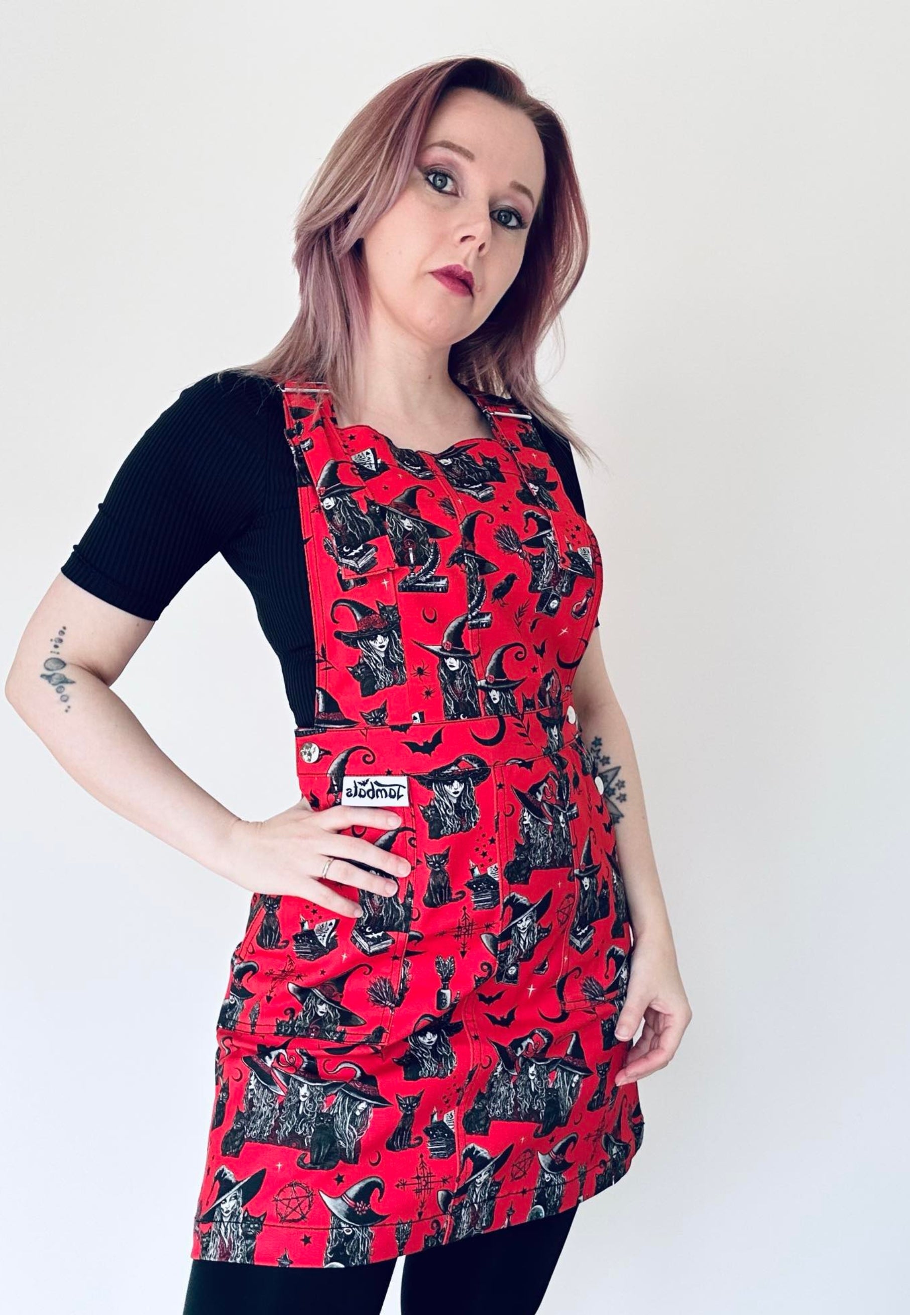A person wearing a red pinafore dress with a witchy print, featuring deep hip pockets and side button fastenings.