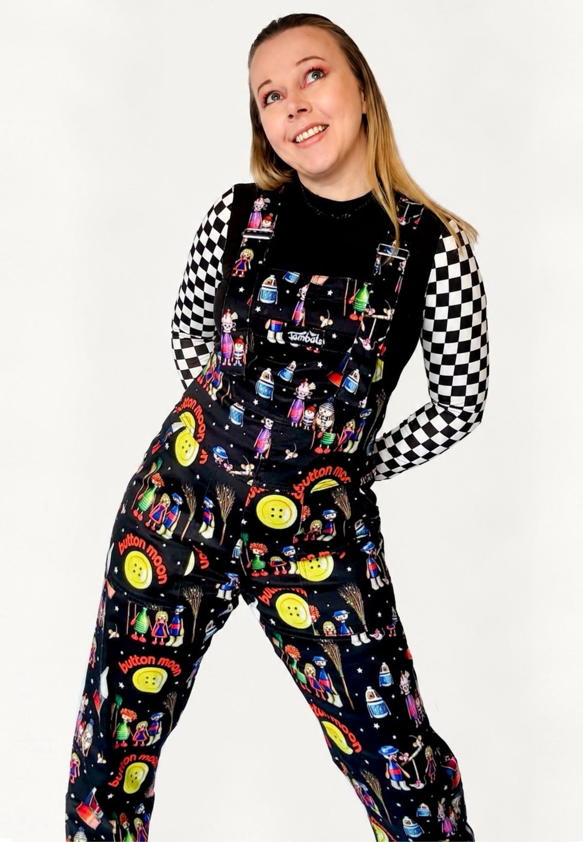 A woman modeling black dungarees with a colorful retro themed print of the 80s children’s tv show characters from Button Moon, worn over a black and white checkered long sleeve shirt against a plain white background.